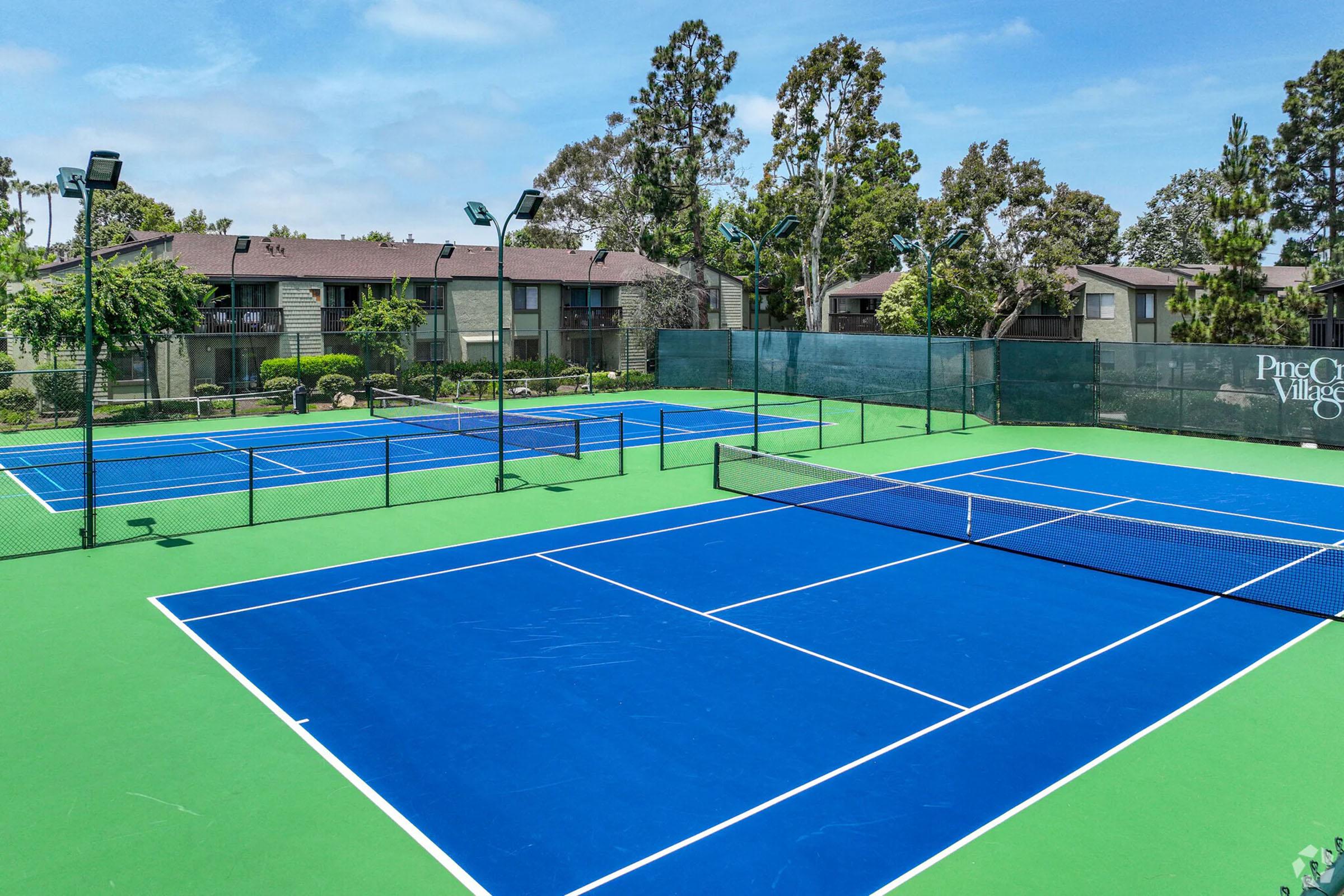 Two well-maintained tennis courts with blue surfaces and green boundaries, surrounded by trees and apartment buildings. The courts feature nets, fence barriers, and overhead lighting, under a clear sky.