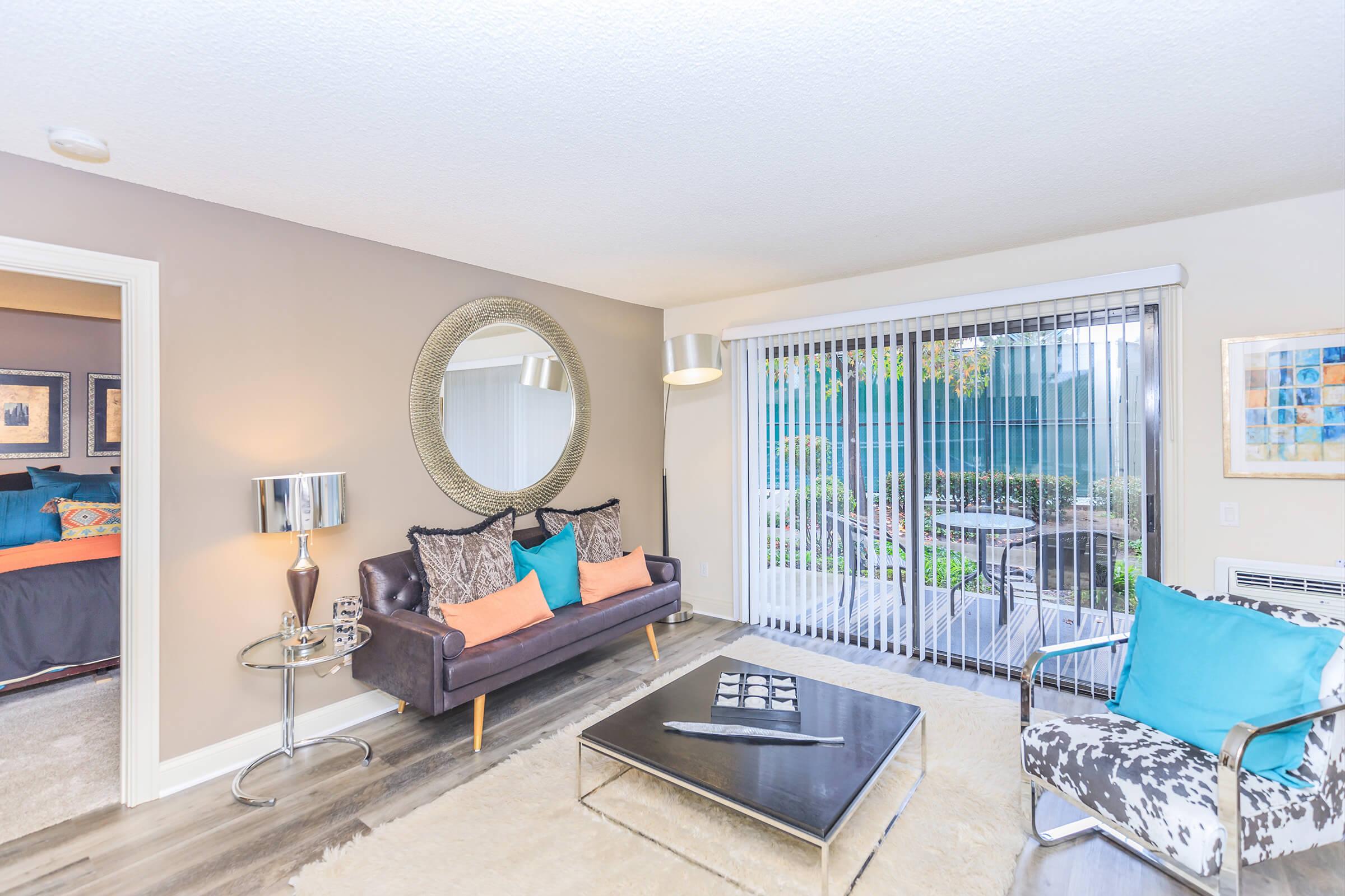A cozy living room featuring a dark sofa with colorful pillows, a round mirror on the wall, a sleek coffee table, and modern decor. Large sliding glass doors provide access to an outdoor area, complemented by a stylish floor lamp and accent chair. The walls are painted a soft neutral color.