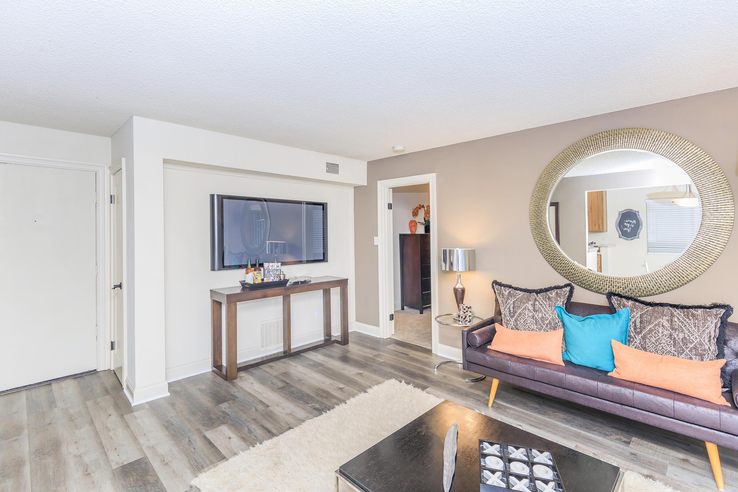 A modern living room featuring a dark leather couch with colorful cushions, a wooden coffee table, and a decorative rug. A flat-screen TV is mounted on the wall above a console table, with a large round mirror and contemporary decor elements visible. A doorway leads to another space.