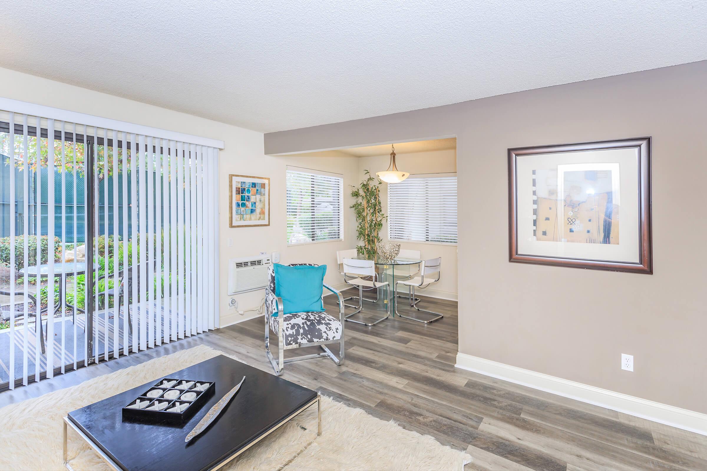 Cozy living space featuring a stylish black coffee table with decorative items, a chic armchair in teal and white, and a dining area with a glass table and chairs. Large windows with blinds allow natural light to brighten the room, while a sliding glass door leads to an outdoor patio with greenery.