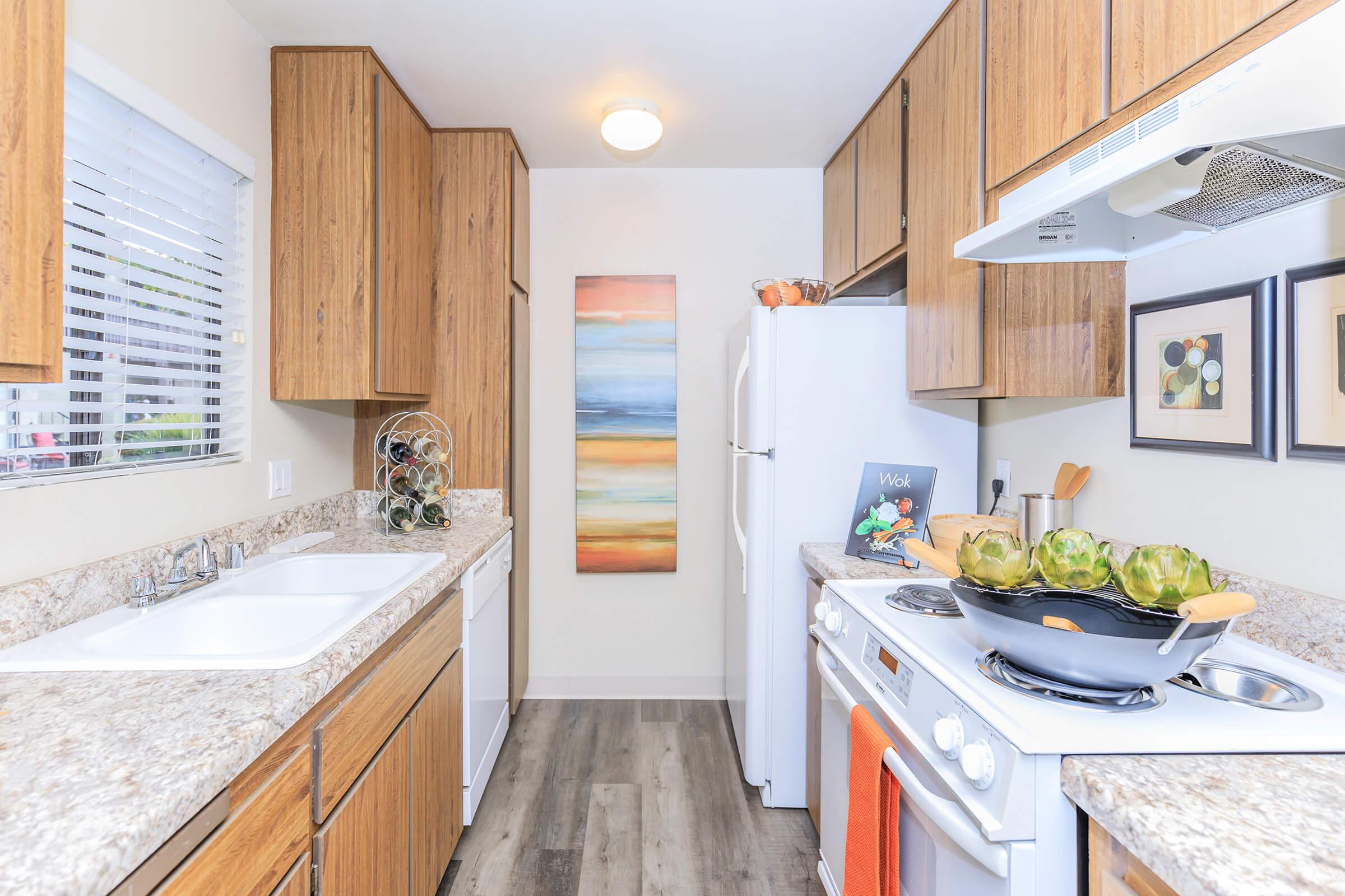 A modern kitchen featuring wooden cabinets, a gray granite countertop, and a white refrigerator. The sink is situated next to the stove, with a large frying pan on the stovetop. A colorful abstract painting hangs on the wall, and there are decorative items and plants on the counter, creating a warm and inviting atmosphere.