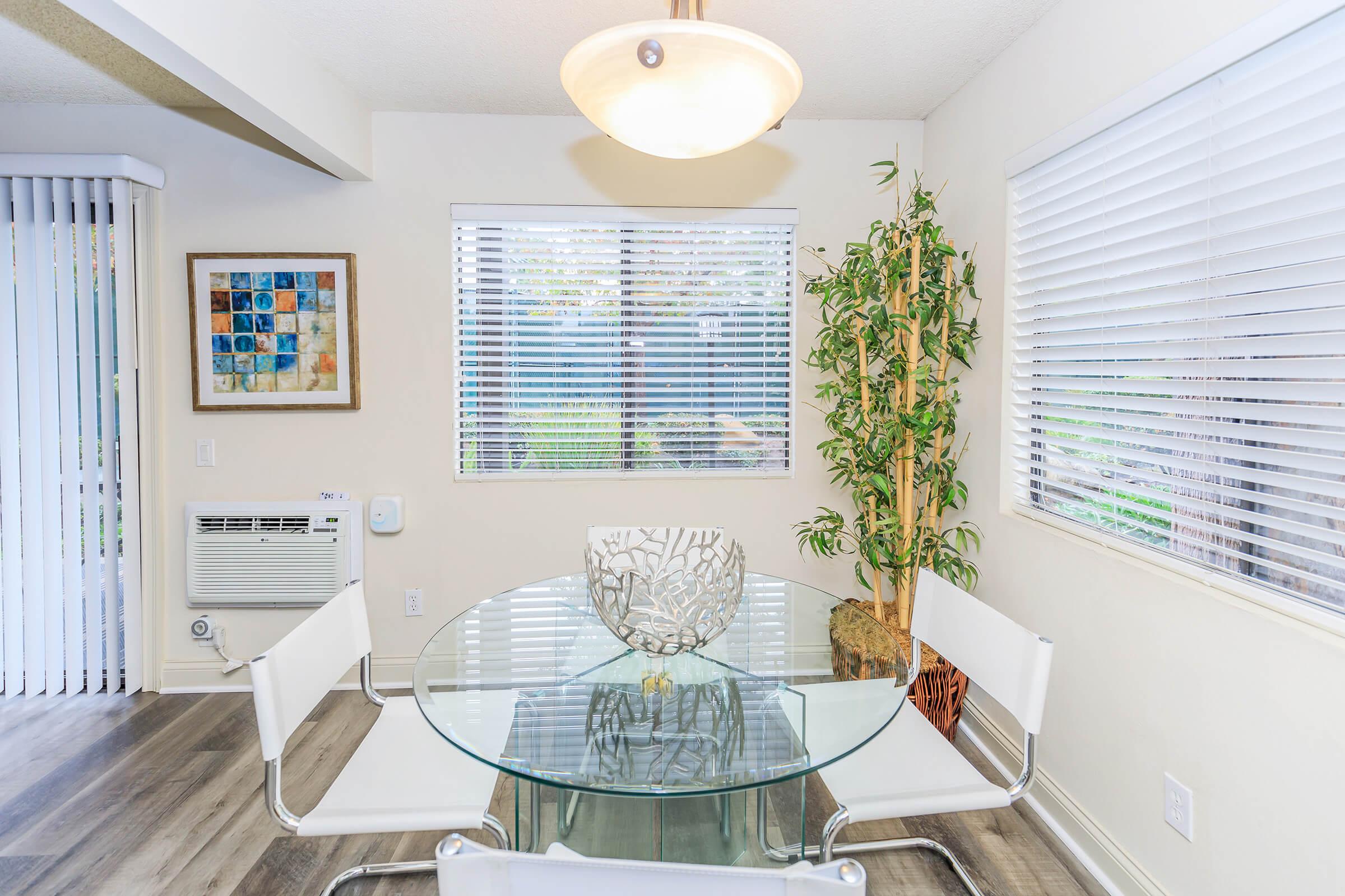 A bright dining area featuring a round glass table with a decorative bowl at the center, surrounded by four white chairs. There’s a potted green plant in the corner, and a large window with blinds allows natural light to fill the space. An air conditioning unit is visible along with a framed artwork on the wall.