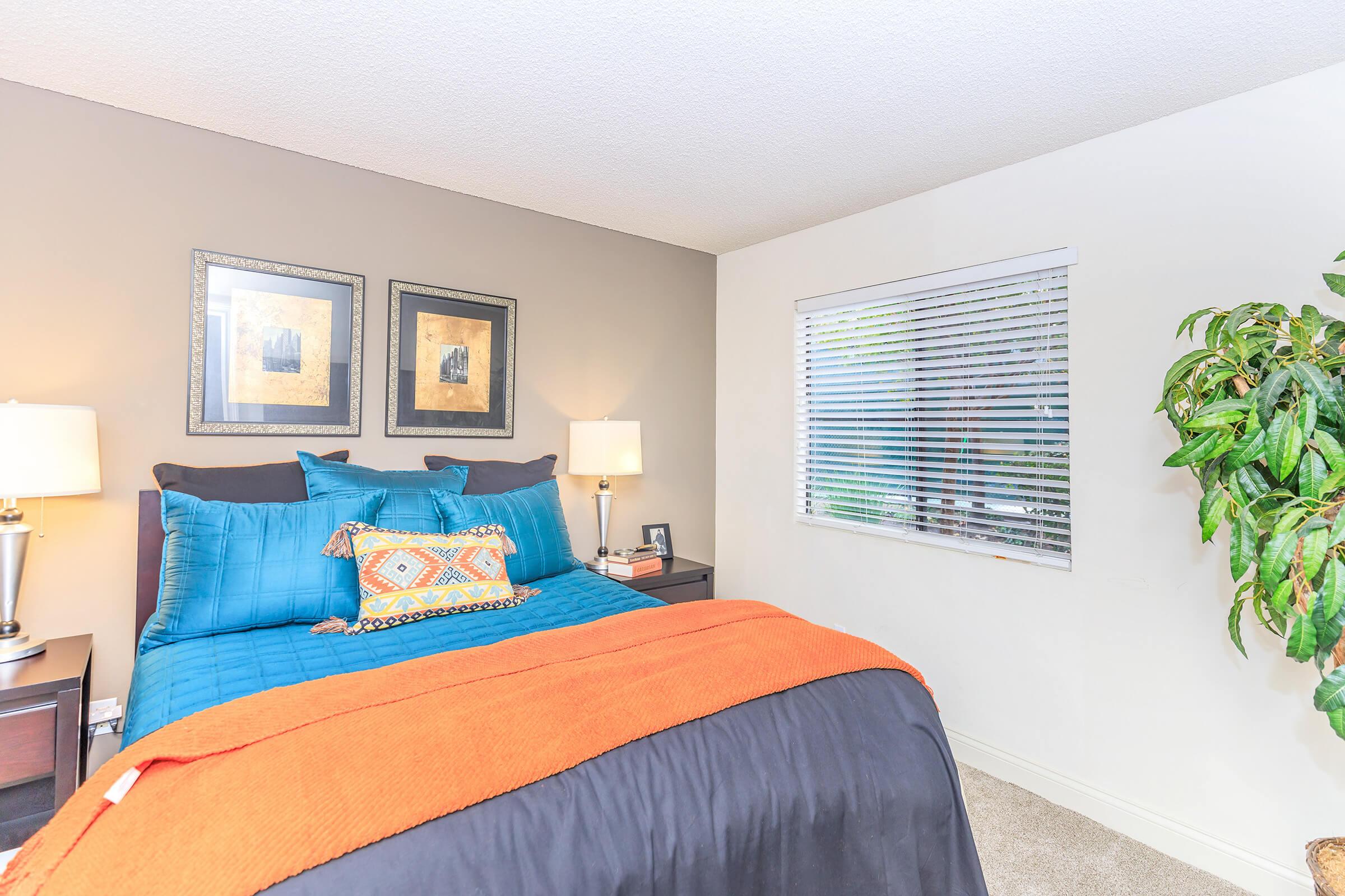 A cozy bedroom featuring a bed with a turquoise and black comforter, an orange throw blanket, and decorative pillows. The walls are painted in a muted tone with two framed artworks. There are bedside lamps on black nightstands, and a window with blinds allows natural light. A green plant adds a touch of nature.