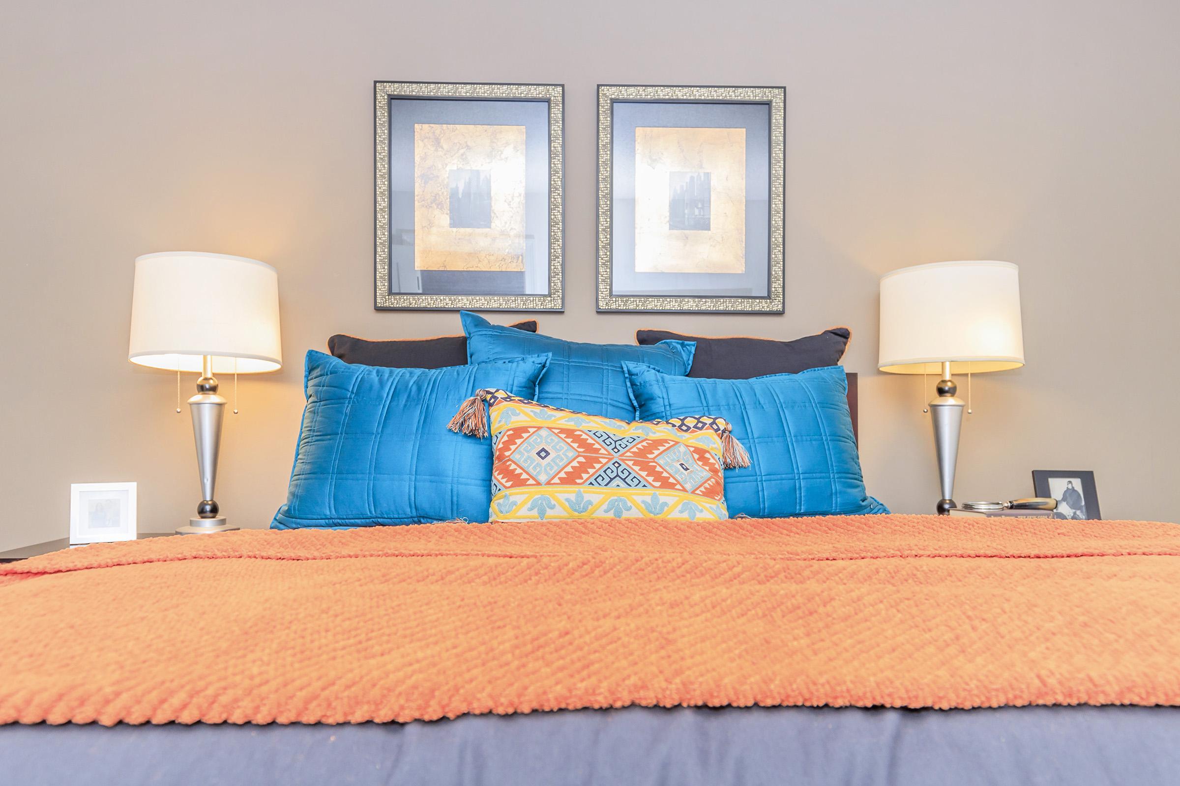 A cozy bedroom setup featuring a neatly made bed with blue and orange bedding. The headboard is adorned with two framed artworks. On either side of the bed, there are matching lamps providing soft lighting. A decorative pillow with geometric patterns adds a pop of color, while a small photo frame sits on one side.