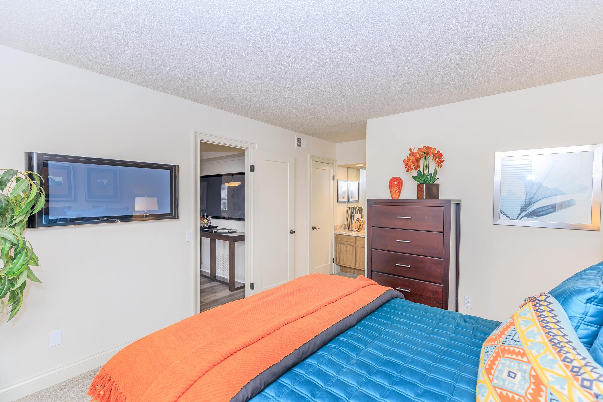 A cozy bedroom featuring a blue bedspread with colorful pillows, an orange throw blanket, and a wooden dresser. A flat-screen TV is mounted on the wall opposite the bed. The doorway leads to a bright, modern bathroom. Decorative plants and an orange vase with flowers add warmth to the space.