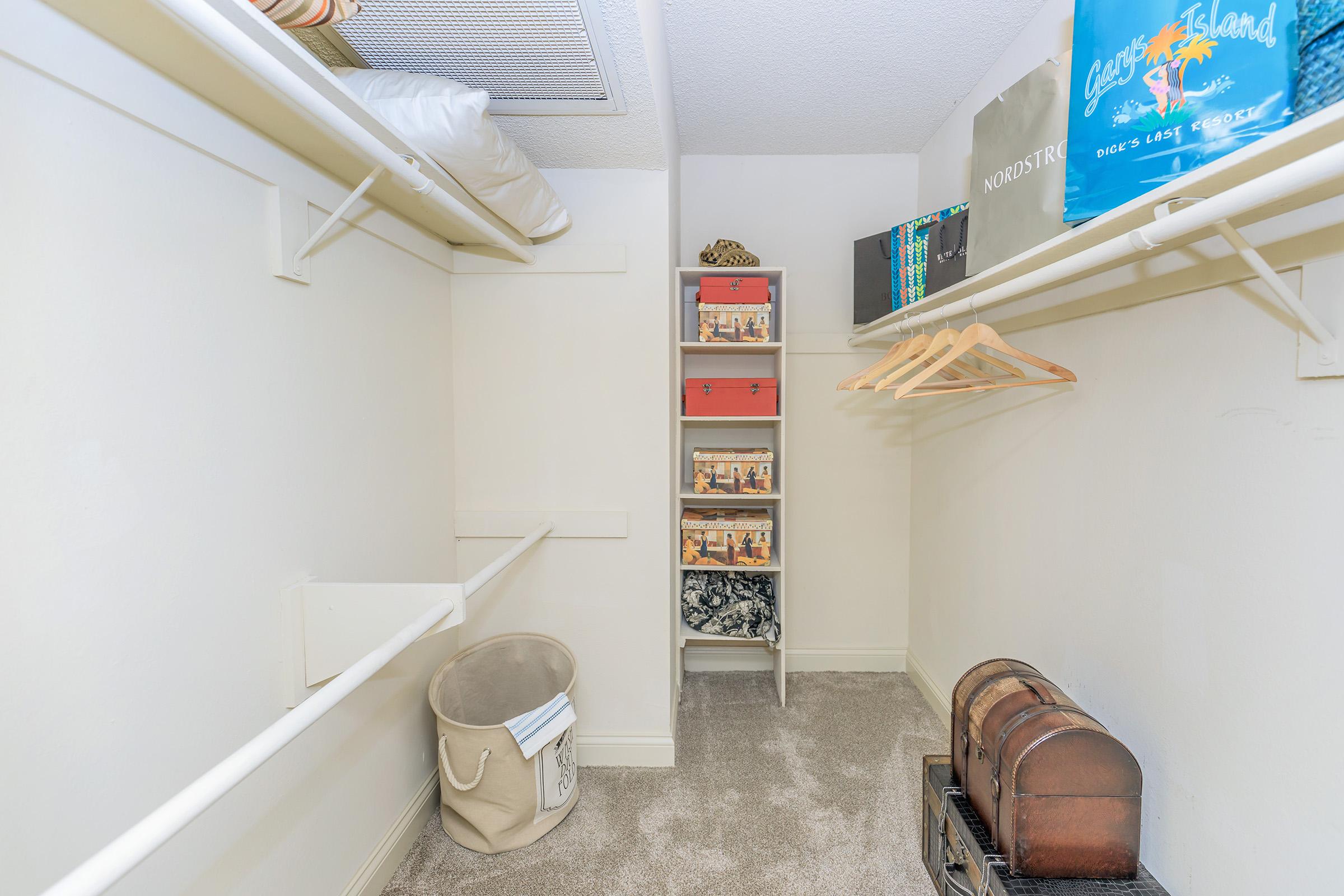 A well-organized closet featuring white shelves, a small storage tower with colorful boxes, a bucket, and a vintage chest. The floor is carpeted, and there are hangers on the wall, indicating ample storage space. Bright lighting highlights the neat arrangement.