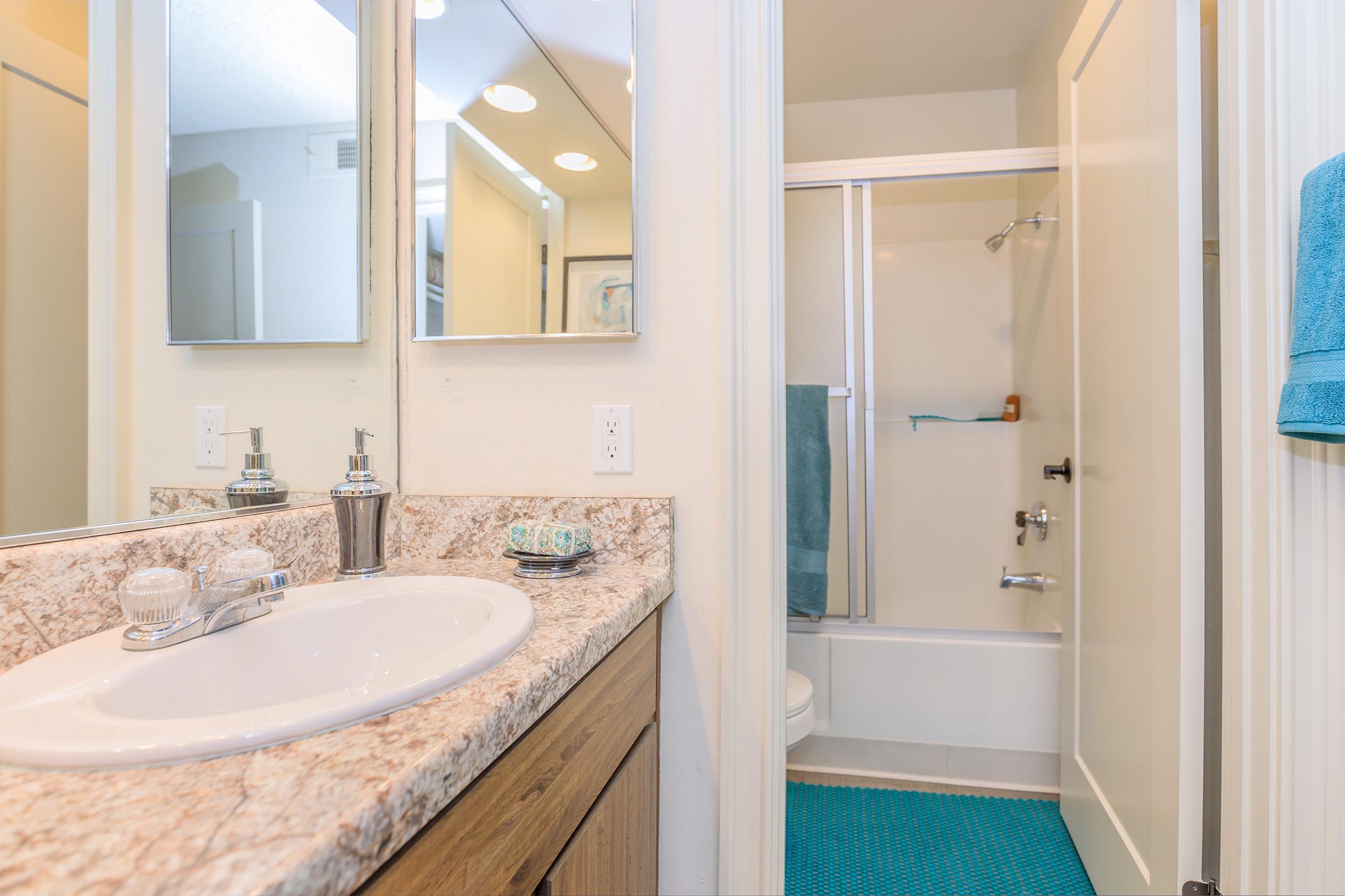 A clean modern bathroom featuring a large mirror, a white sink with silver fixtures, and a countertop with decorative items. The shower area has a glass door and is equipped with a blue towel hanging. The floor is tiled with a light color, and the walls are painted a soft shade, creating a spacious feel.