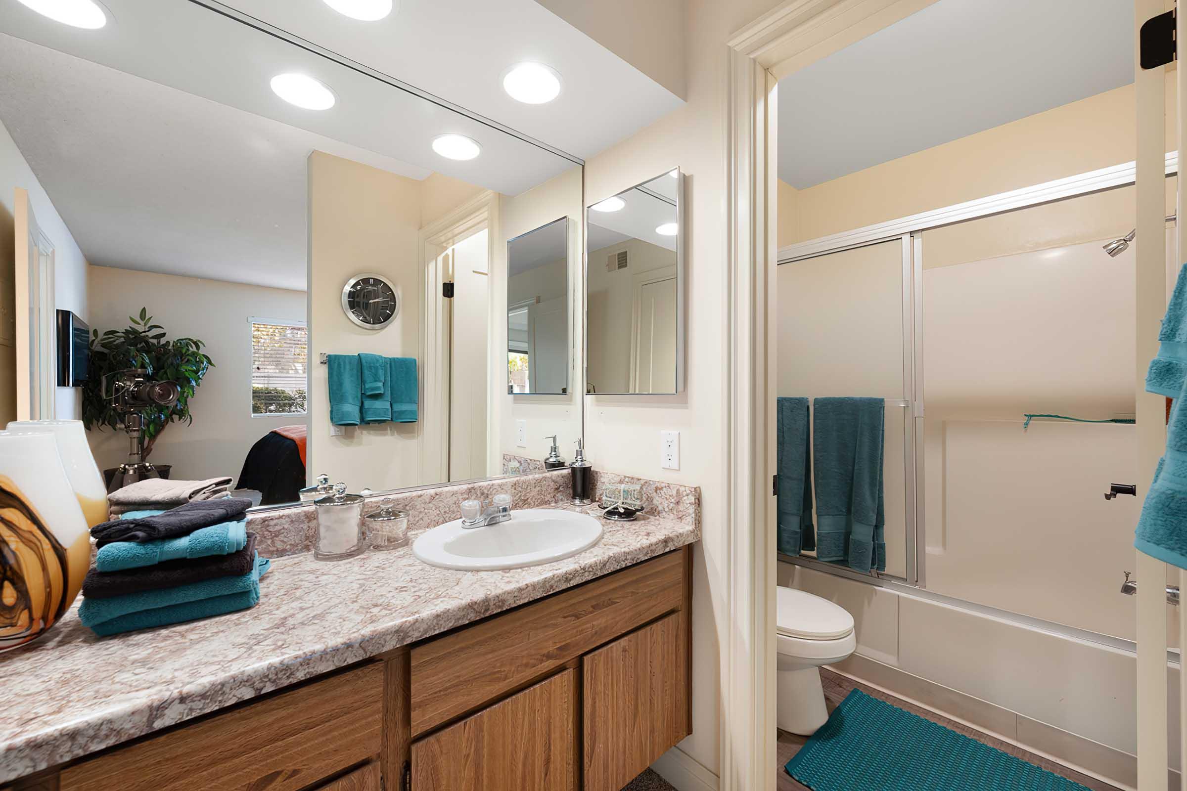 A bright and clean bathroom featuring a double sink vanity with a speckled countertop, two mirrors, and modern lighting. There's a shower with a clear door, a toilet, and teal towels hanging nearby. A potted plant adds a touch of greenery, and a small clock is mounted on the wall.