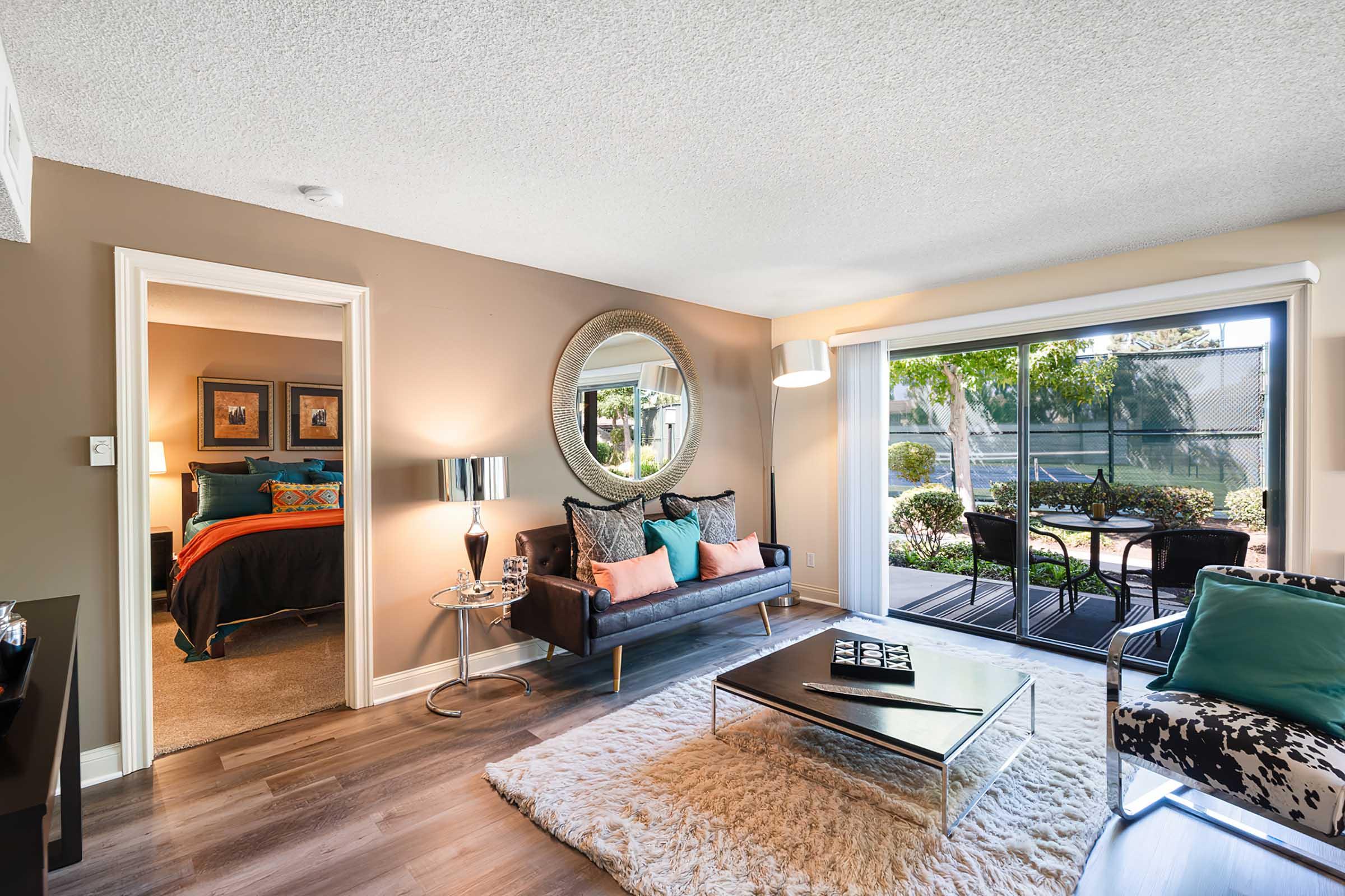 Modern living room featuring a stylish black sofa with decorative pillows, a round mirror on the wall, and a sleek coffee table. A large window provides natural light and overlooks an outdoor patio with a dining area. The room has warm, neutral tones and a cozy area rug.