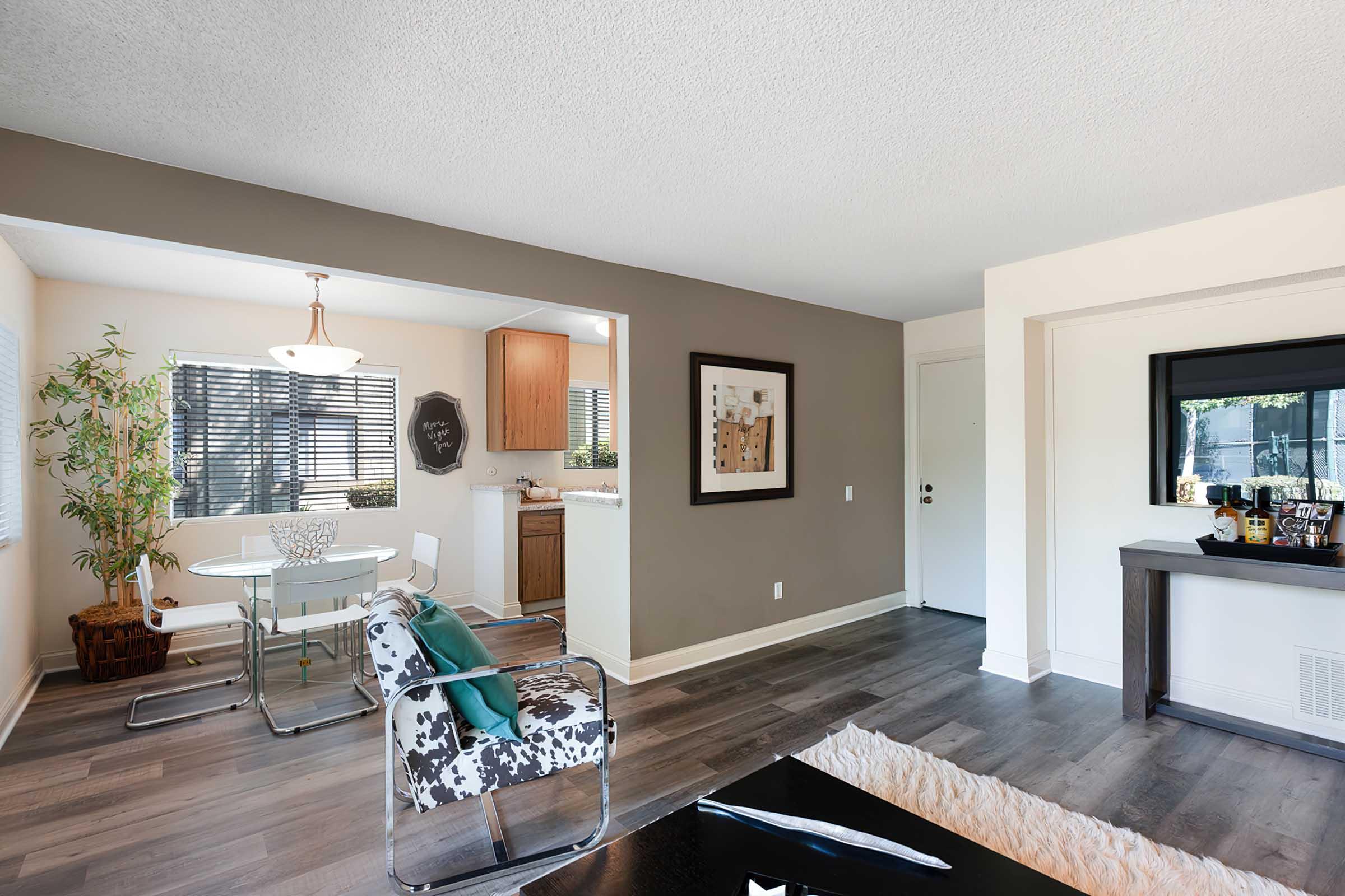 A modern living space featuring a dining area with a glass table and stylish chairs, a kitchen area visible in the background, and an inviting living room with hardwood-style flooring. There's a decorative black-framed artwork and a small bar setup on a console table. Natural light brightens the space.