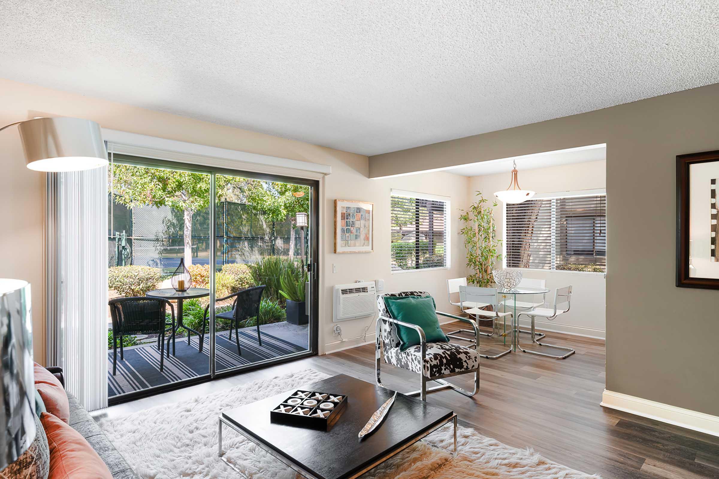 A modern living room featuring large glass doors that open to a garden view. Inside, there is a cozy seating area with a plush rug, a stylish coffee table, and contemporary furniture. A dining space with a glass table is visible, accompanied by natural light and greenery outside.