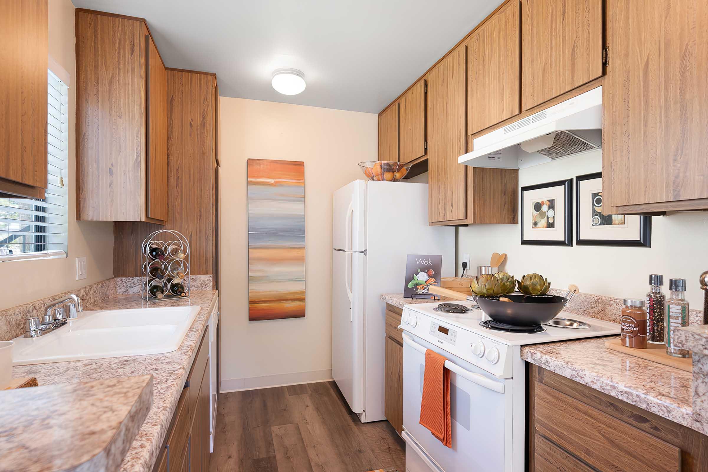 A modern kitchen featuring wooden cabinets, a white refrigerator, and a stove with an orange towel. The countertop is adorned with decorative items and a bowl of greenery. There's a wine rack on the wall and artwork hanging near the window, creating a warm and inviting atmosphere.