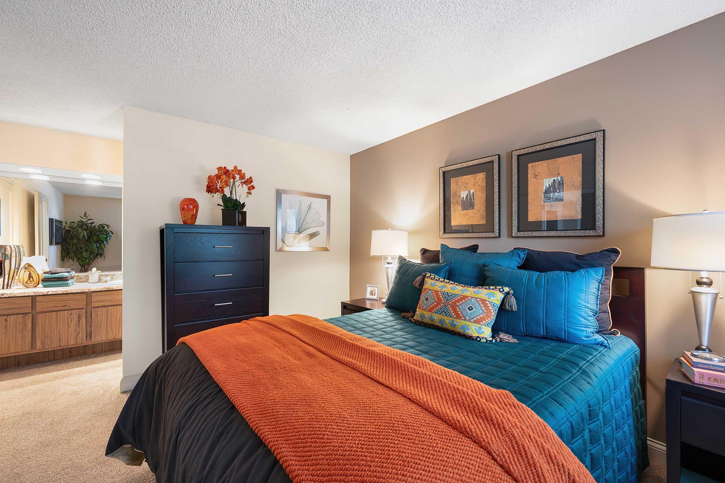 Cozy bedroom featuring a blue and black quilted bedspread adorned with vibrant pillows. A dark dresser stands beside the bed, complemented by framed artwork on the wall. Soft lighting from bedside lamps creates a warm ambiance, while an orange throw adds a pop of color. A bathroom is visible in the background.