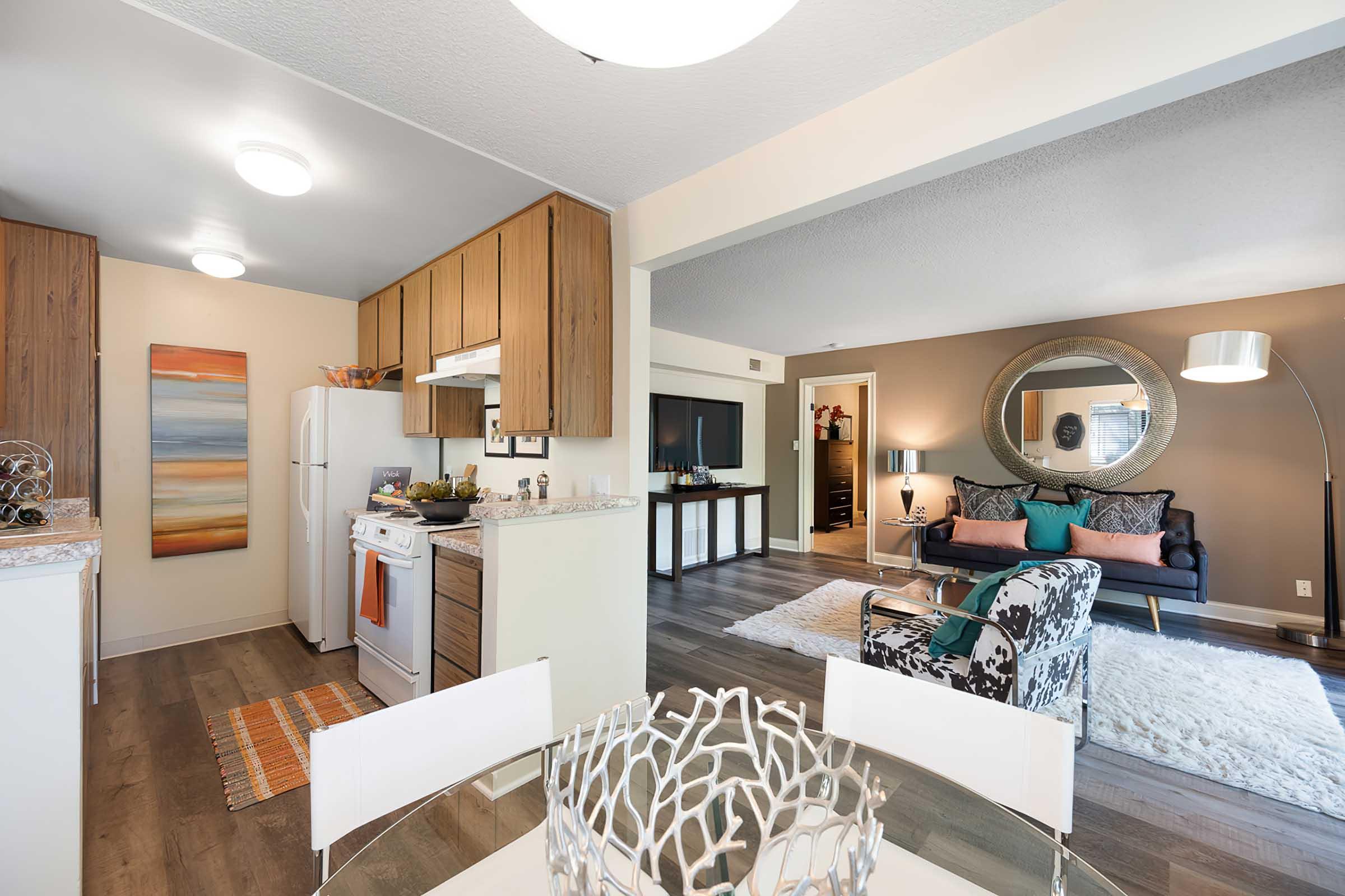 Modern apartment interior showcasing an open layout. The kitchen features wood cabinets and white appliances, while the living area has a stylish sofa, decorative throw pillows, and a round mirror. A dining table with a unique centerpiece sits in the foreground, and the space is illuminated by soft lighting.