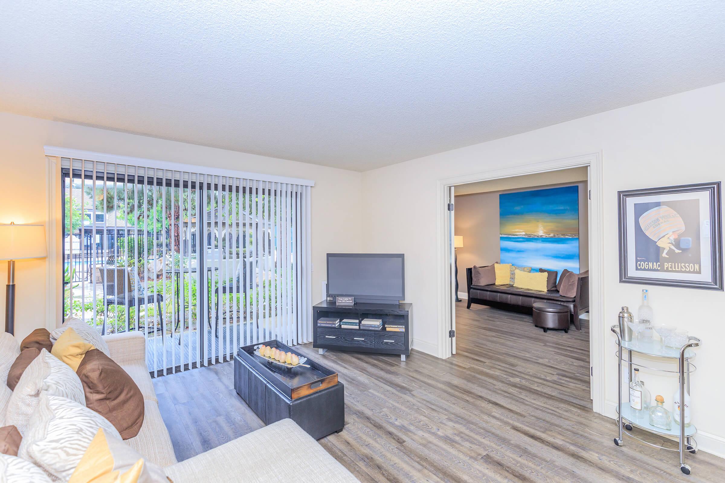 Bright living room featuring a beige sofa with decorative pillows, a black coffee table with a tray, and a television on a black media console. Large sliding glass doors provide natural light and open to a view of greenery outside. A second inviting living space is visible through an open doorway, adorned with artwork.