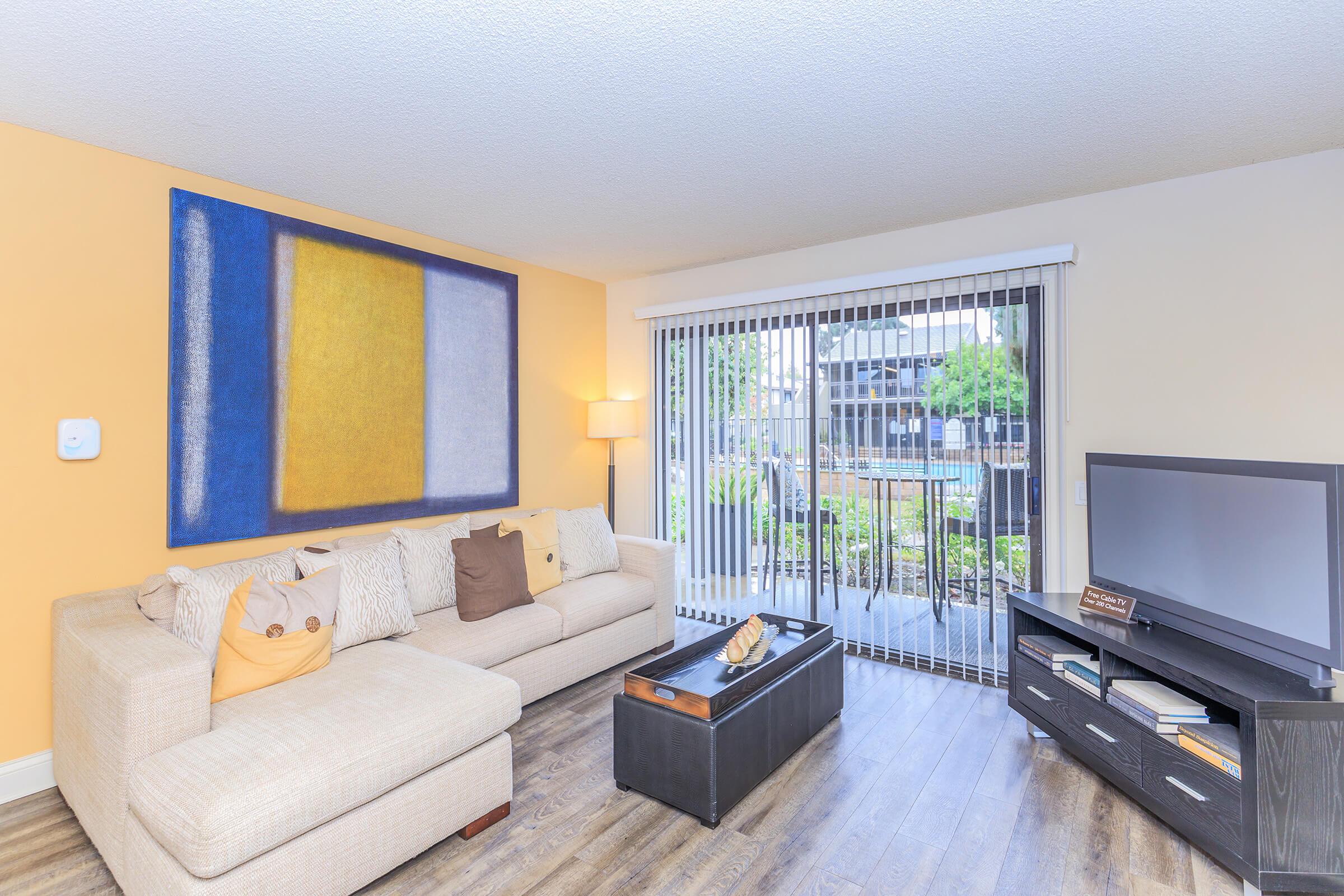 A cozy living room featuring a light-colored sectional sofa adorned with decorative pillows, a modern coffee table, and a flat-screen TV. The wall behind the sofa showcases a large abstract painting, and a sliding glass door leads to a patio with greenery outside.