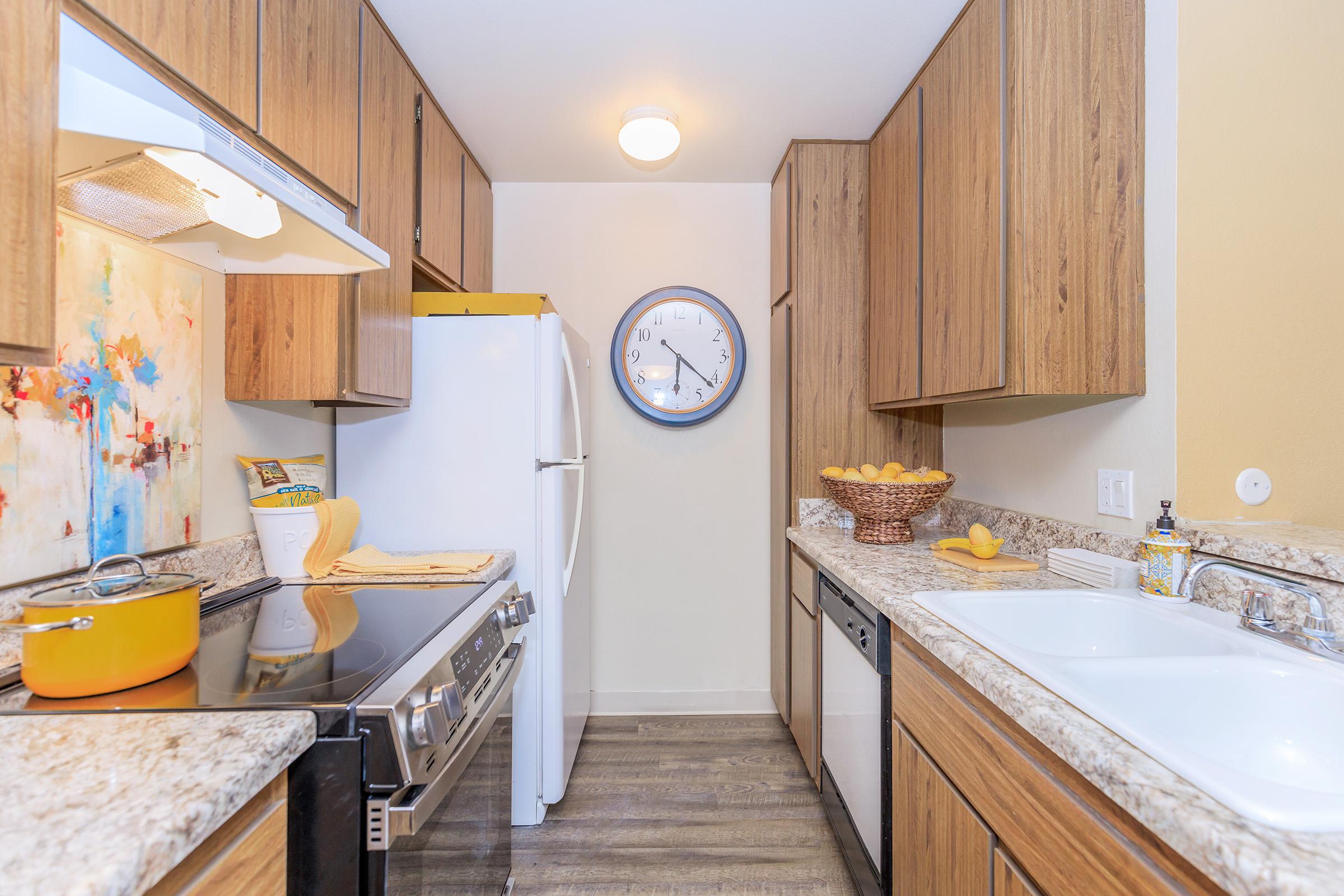 A modern kitchen featuring wooden cabinetry, granite countertops, and stainless steel appliances. The space includes a white refrigerator, a yellow decorative bowl with lemons, a clock on the wall, and a stovetop. Natural light brightens the room, enhancing its inviting atmosphere.