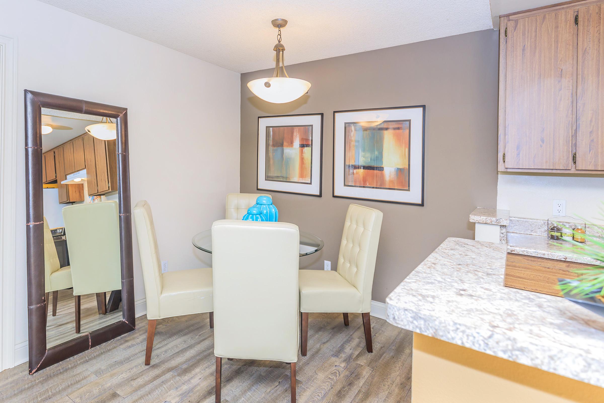 A modern dining area featuring a round glass table surrounded by four light-colored upholstered chairs. A large mirror reflects the space, and two framed abstract paintings adorn the wall. A pendant light hangs above the table, adding warmth to the neutral palette with wood accents.