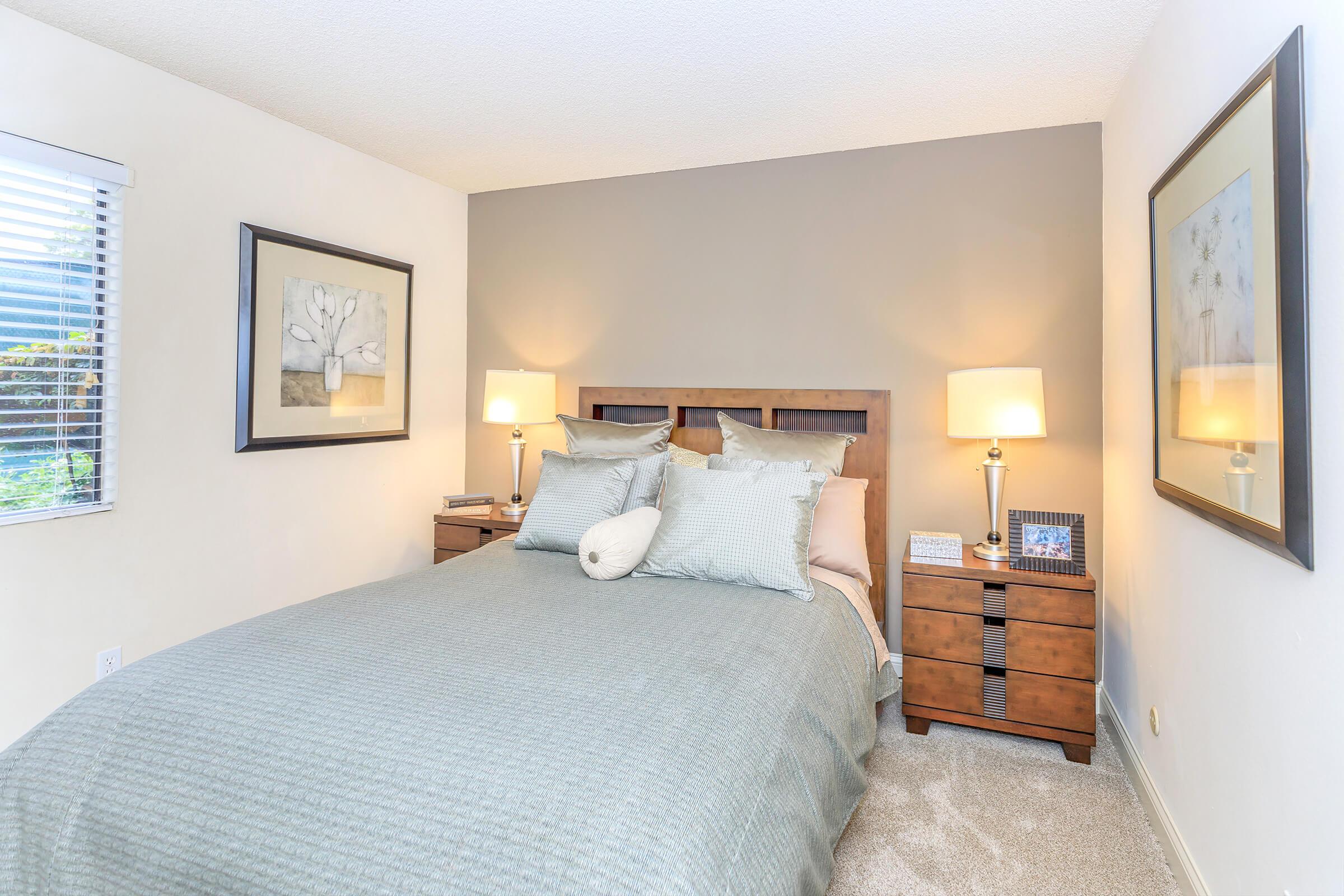 A cozy bedroom featuring a queen-sized bed with a light blue comforter and decorative pillows. Beside the bed, there are two wooden nightstands with lamps, and framed artwork is displayed on the wall. The room has neutral-colored walls and a carpeted floor, with natural light coming through a window.