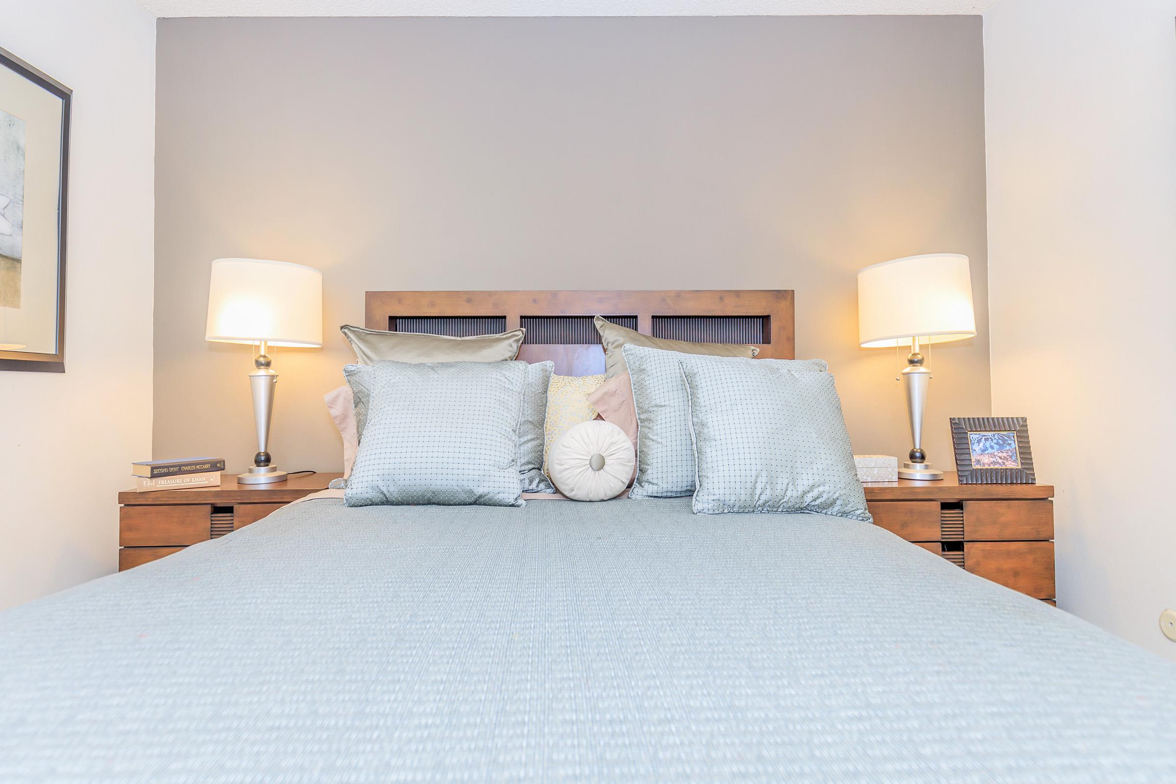 A cozy bedroom featuring a neatly made bed with decorative pillows, two bedside lamps casting a warm glow, and a wooden headboard. A framed picture and a book are placed on the nightstands beside the bed, against a neutral-colored wall.