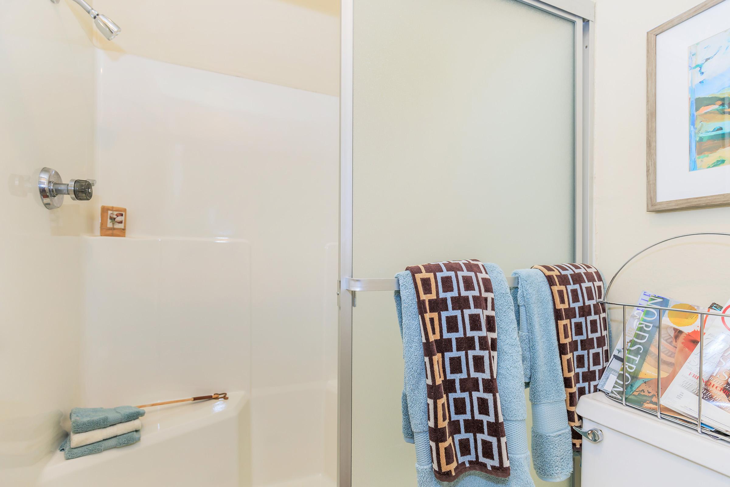 A clean bathroom featuring a shower with a glass door, two neatly hung towels in brown and blue, a small wooden shelf with toiletries, and a decorative basket on the side filled with items, alongside a colorful framed painting on the wall.