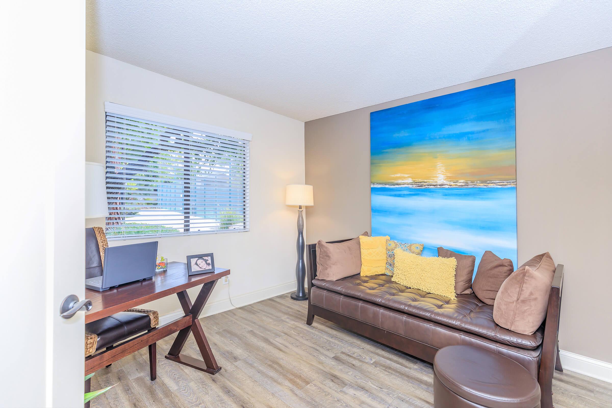 A modern living space featuring a brown leather sofa adorned with yellow and brown cushions, a small desk with a chair, and a lamp. A large painting of a seascape hangs on the wall, and there is natural light coming through a window with blinds. The floor is made of wood laminate.