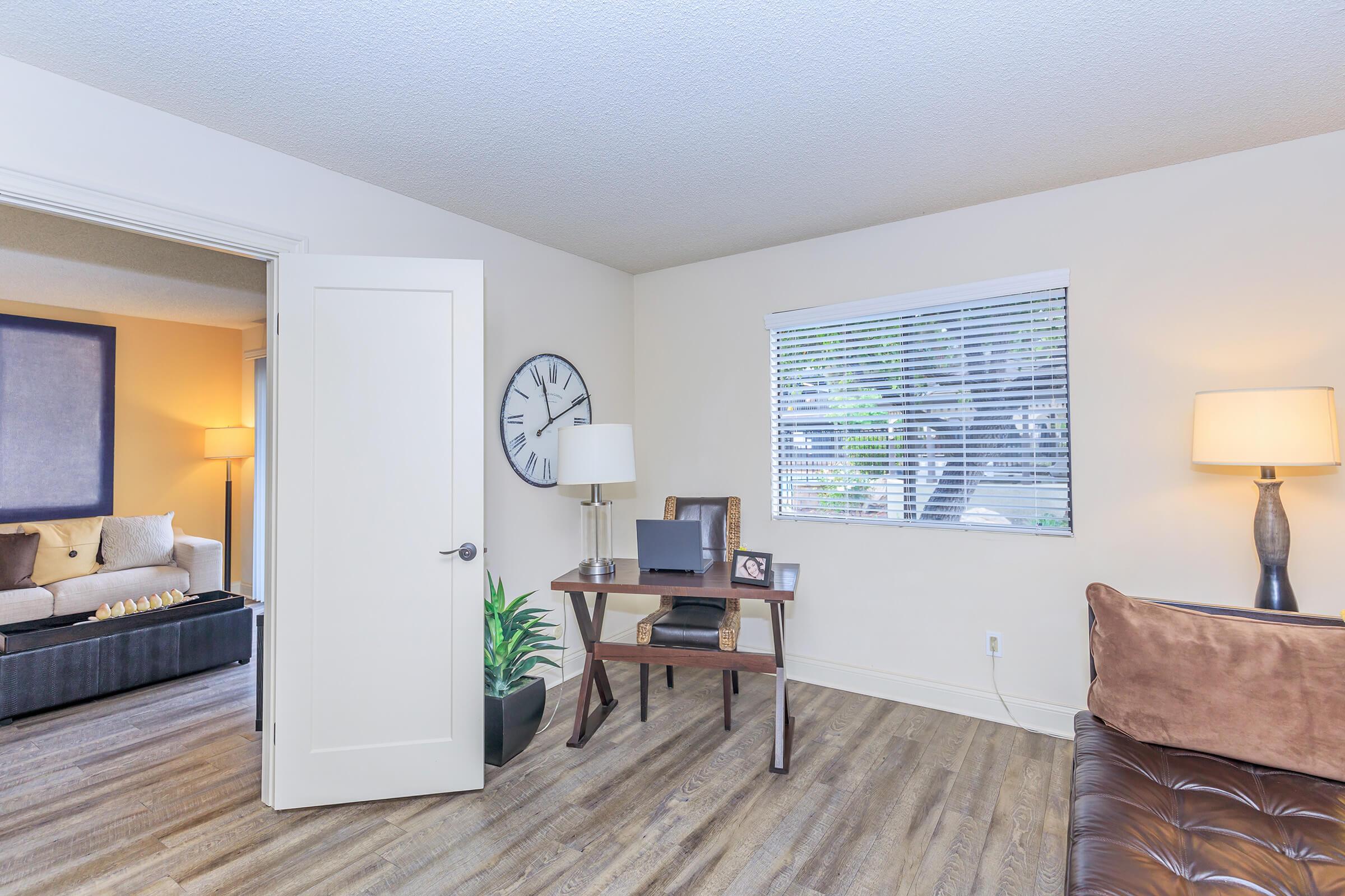 A bright, modern living space featuring a wooden desk with a laptop, a potted plant, and framed pictures. A cozy brown couch is visible on the right, while a light-colored wall with a window overlooks the room. A clock adorns the wall, and a soft lamp adds warm lighting to the atmosphere.