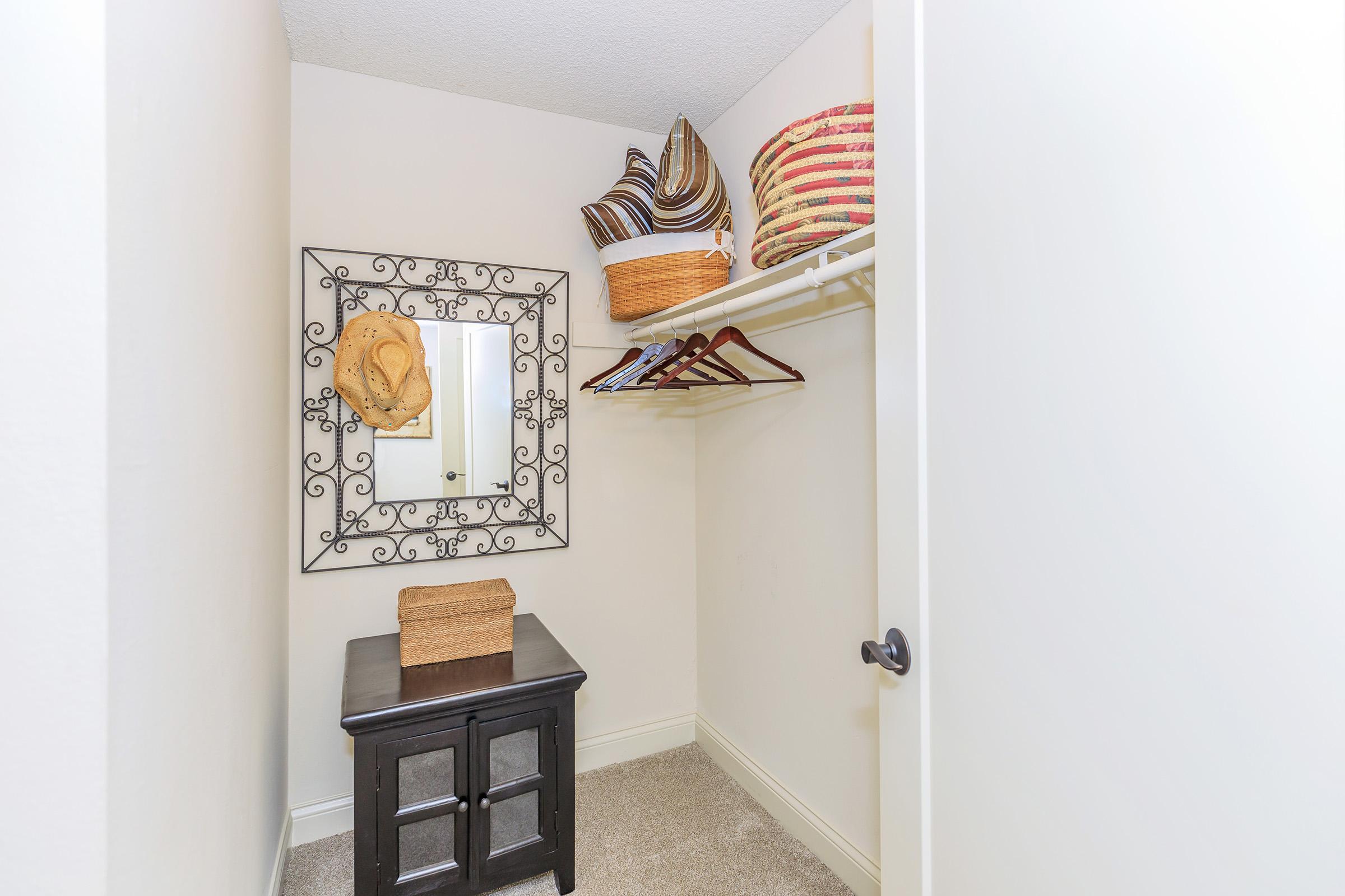 A small, organized closet featuring a full-length mirror with an ornate frame, a black wooden storage table, and a shelf with woven baskets and hangers. Soft beige walls and carpet create a cozy atmosphere.