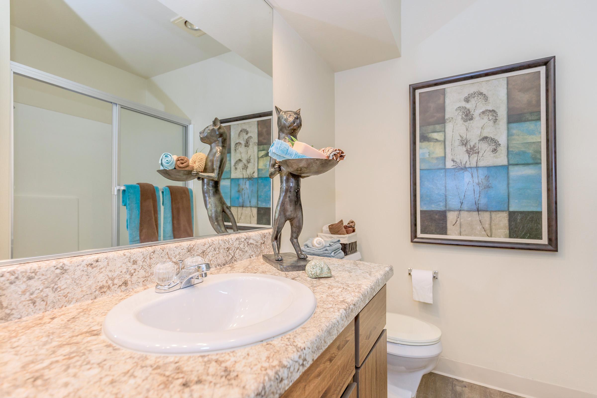 A modern bathroom featuring a countertop sink, a decorative statue of a cat holding towels, and a framed art piece on the wall. The decor includes a soft color palette, towels hanging on a rack, and a toilet in the background, creating a clean and inviting atmosphere.