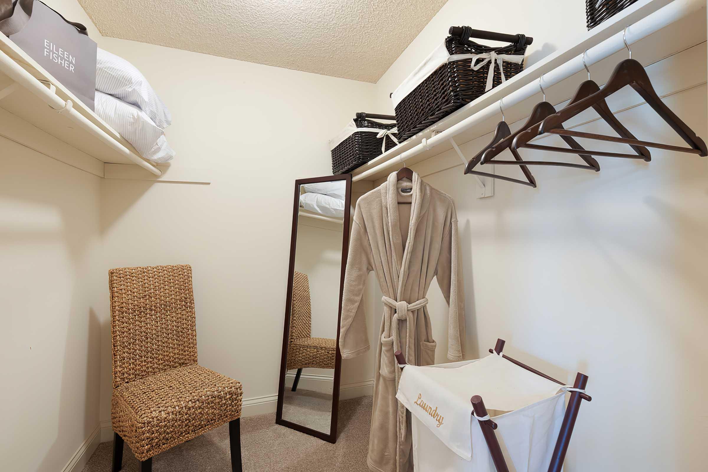 A well-organized closet featuring a plush beige robe hanging on a hook, a full-length mirror, a wicker chair, a laundry basket, and several baskets for storage. The walls are light-colored, and the space is tidy and inviting.