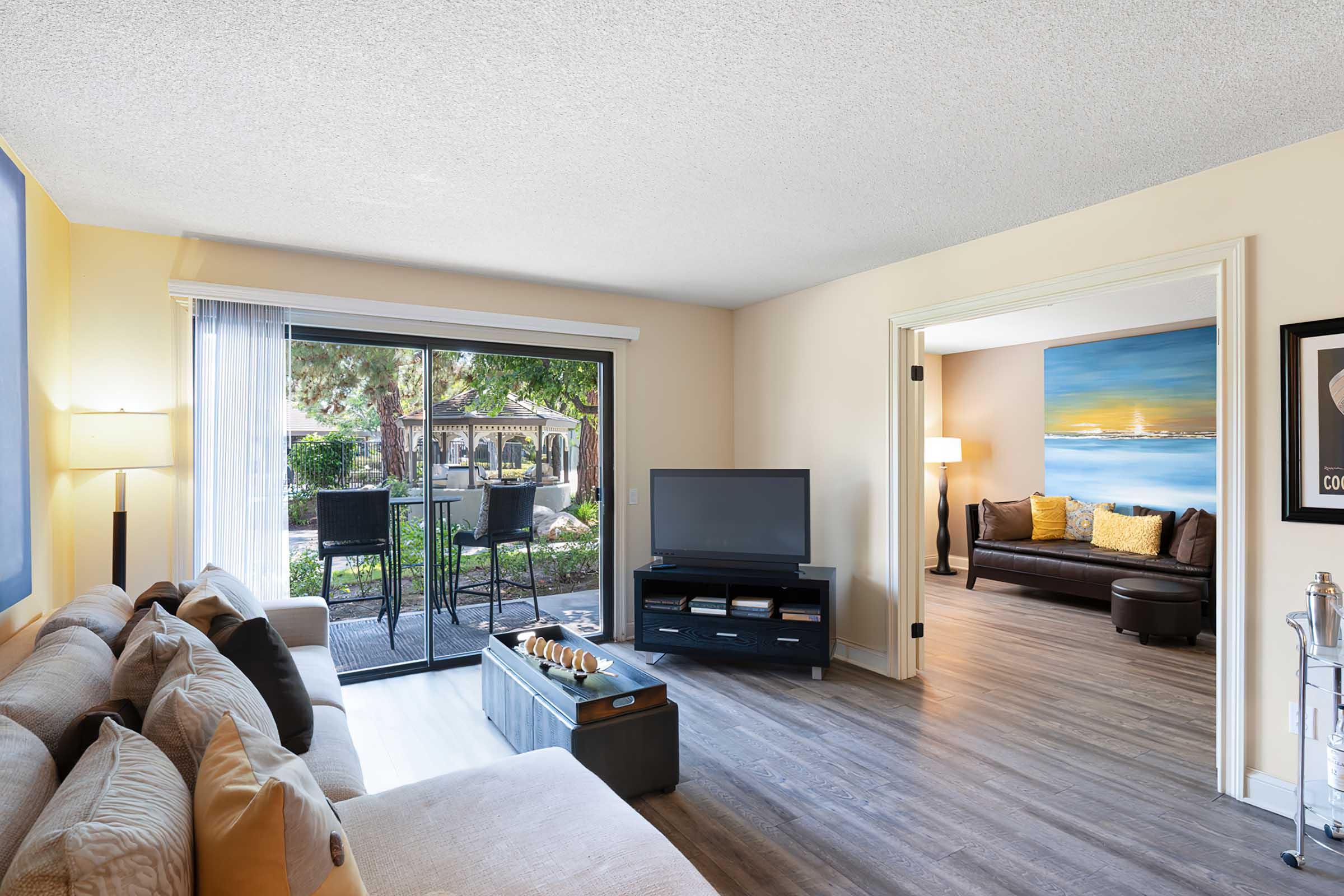 Bright living room with a large window offering a view of the outdoor patio. A comfortable beige sofa and a coffee table with decorative items are situated in the foreground. A TV is on a stand, while a doorway leads to another room featuring a brown sofa and a colorful painting on the wall.