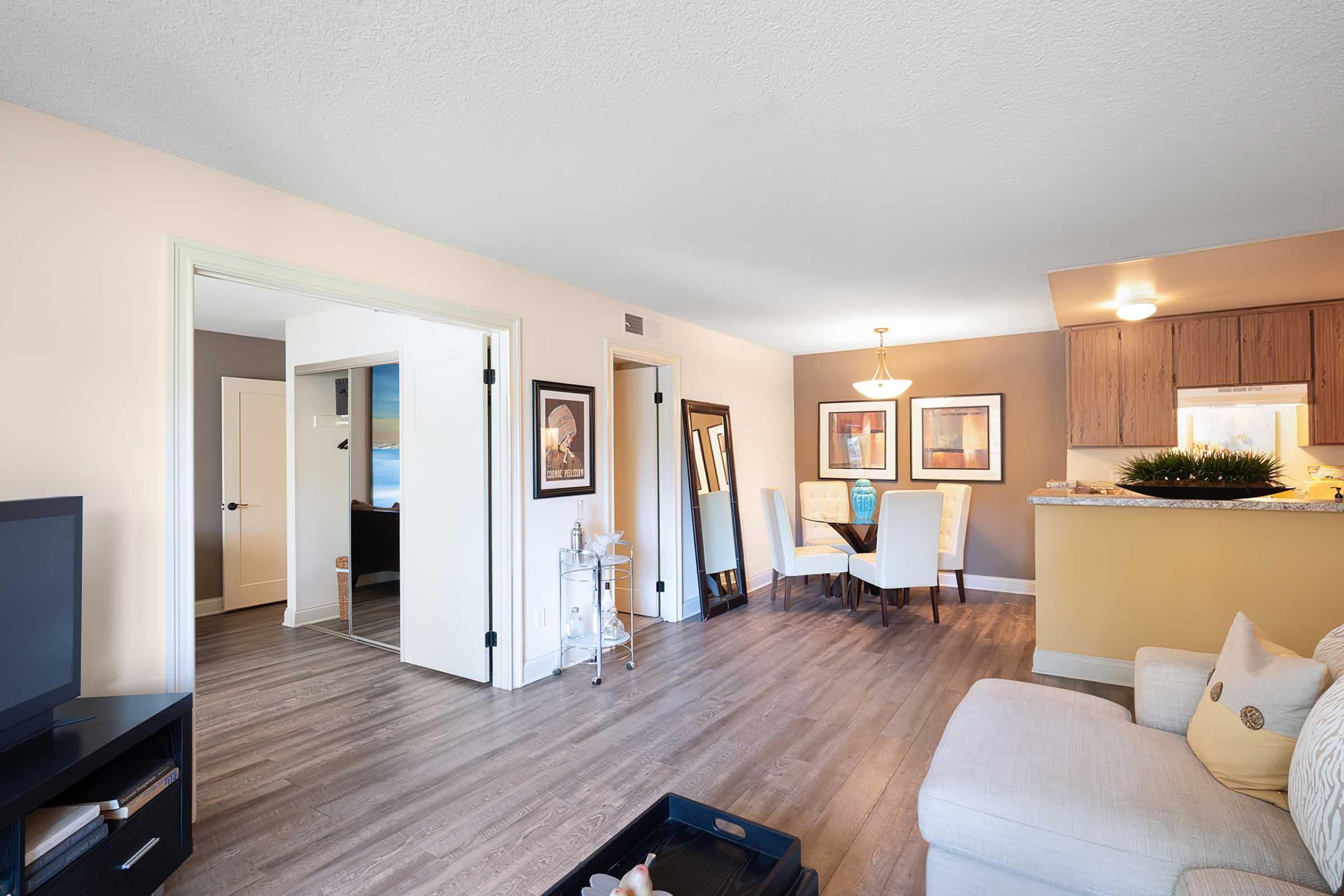 Bright and spacious living area featuring a light-colored couch, a coffee table with decorative items, and a TV. The room opens to a dining area with a round table and chairs. Light walls and modern artwork create a warm atmosphere, while the kitchen is visible in the background, enhancing the open floor plan.