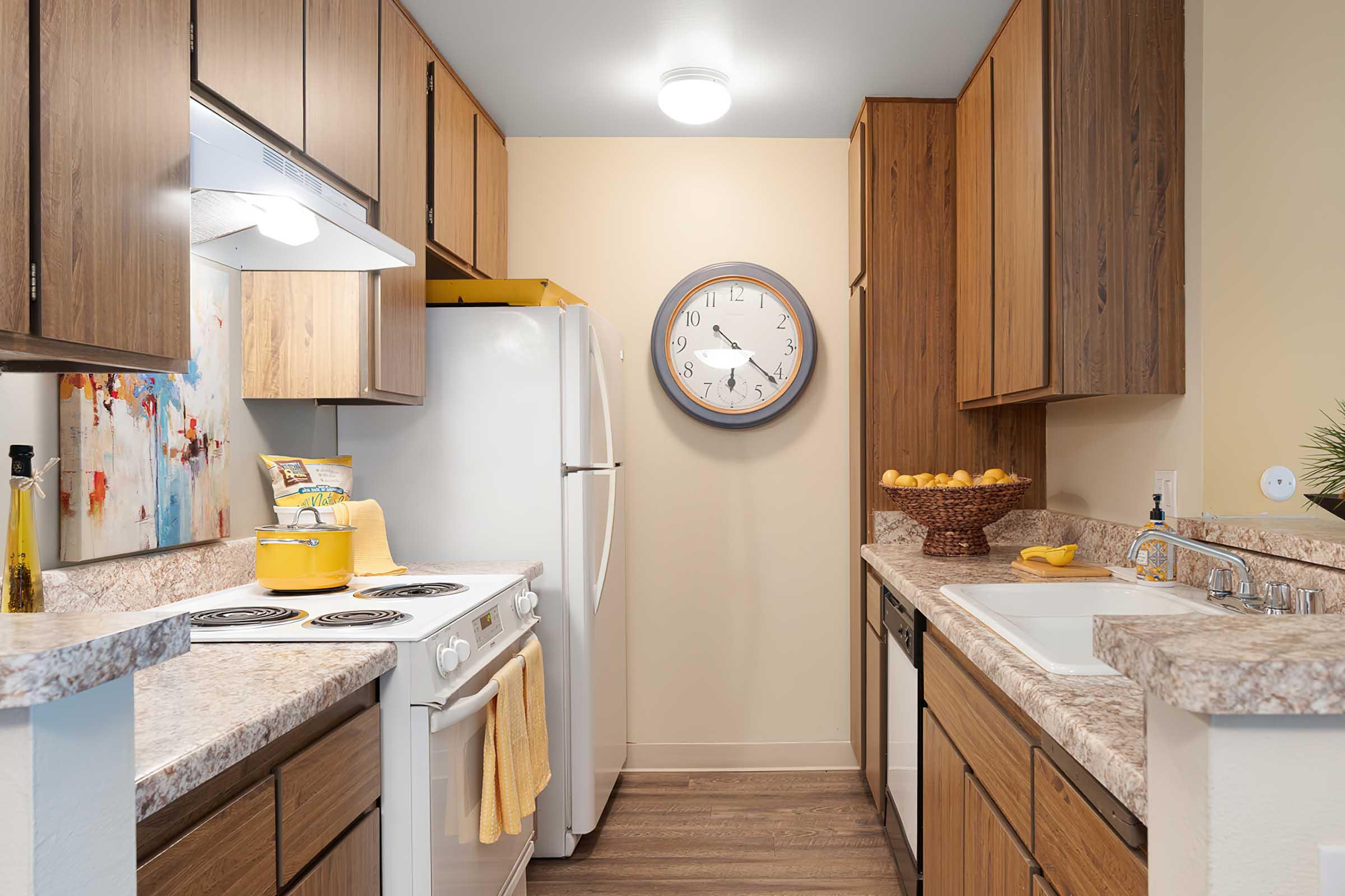 A modern kitchen featuring wooden cabinets, a white refrigerator, and a white stove. The countertop is decorated with a yellow dish towel and a bowl of lemons. A large wall clock and a colorful abstract painting add contrast to the neutral-colored walls. Natural light illuminates the space.
