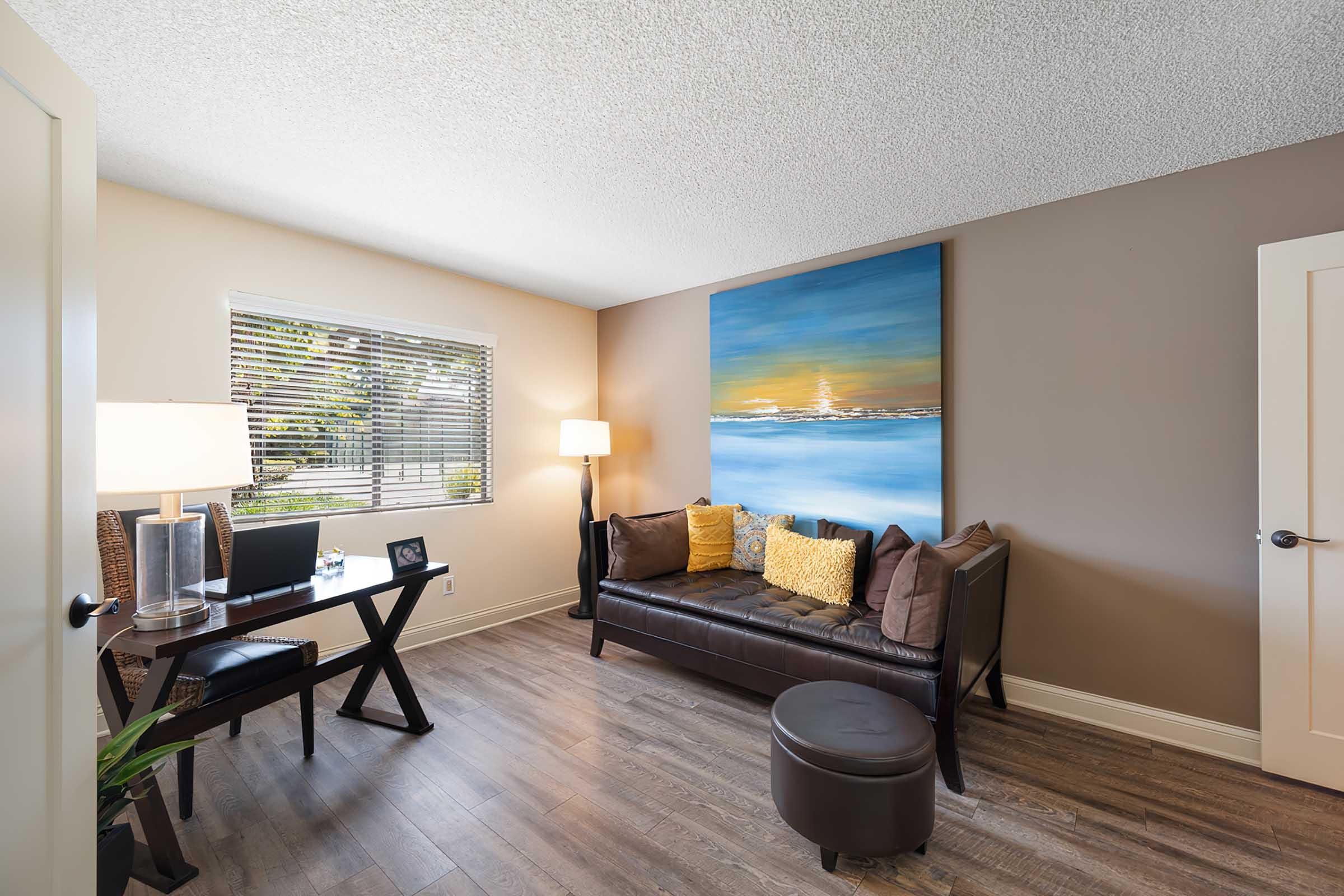 A cozy living space featuring a brown leather couch with yellow pillows, a small round ottoman, a desk with a chair and lamp, and a large abstract painting on the wall. Natural light streams in through a window with blinds, and the room has a warm, inviting atmosphere with wood flooring.