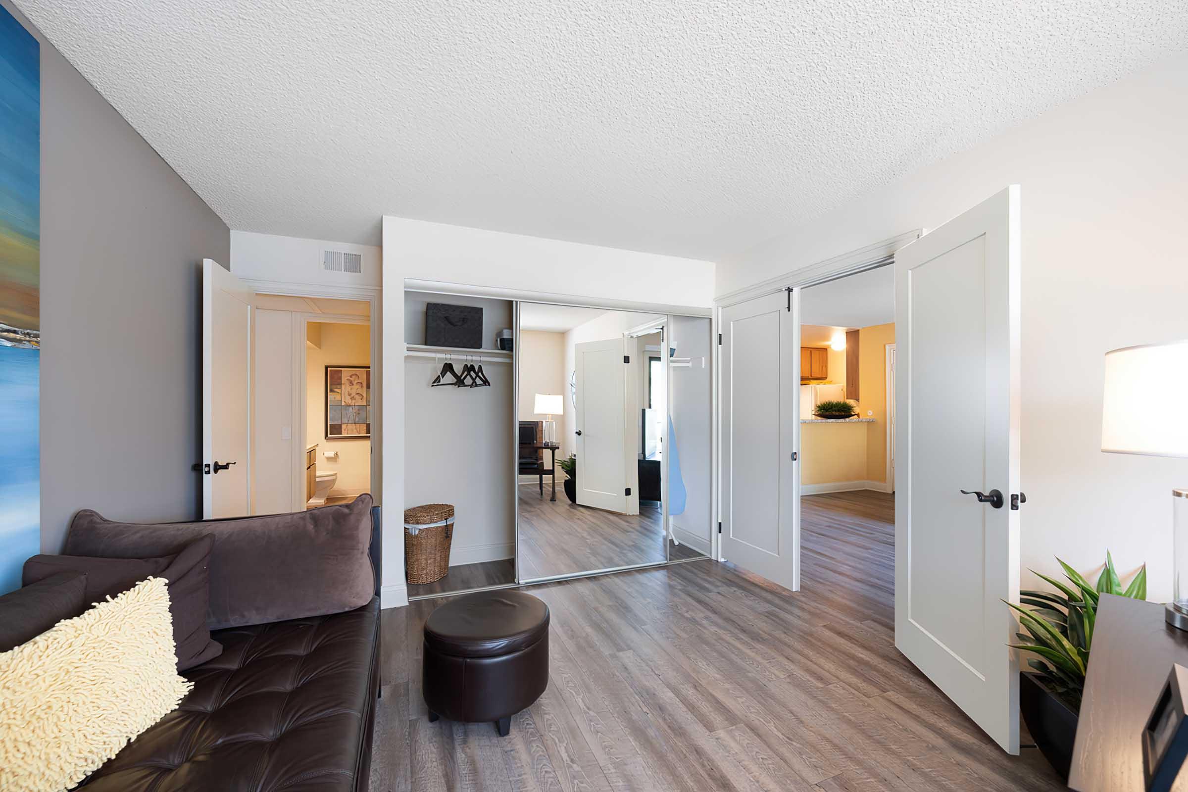 A cozy living space featuring a brown sectional sofa with a decorative pillow, a round ottoman, and a light source from a nearby lamp. The room has an open layout with mirrored closet doors, leading to a bedroom area and a kitchen visible in the background, with wood-like flooring and a plant in the corner.