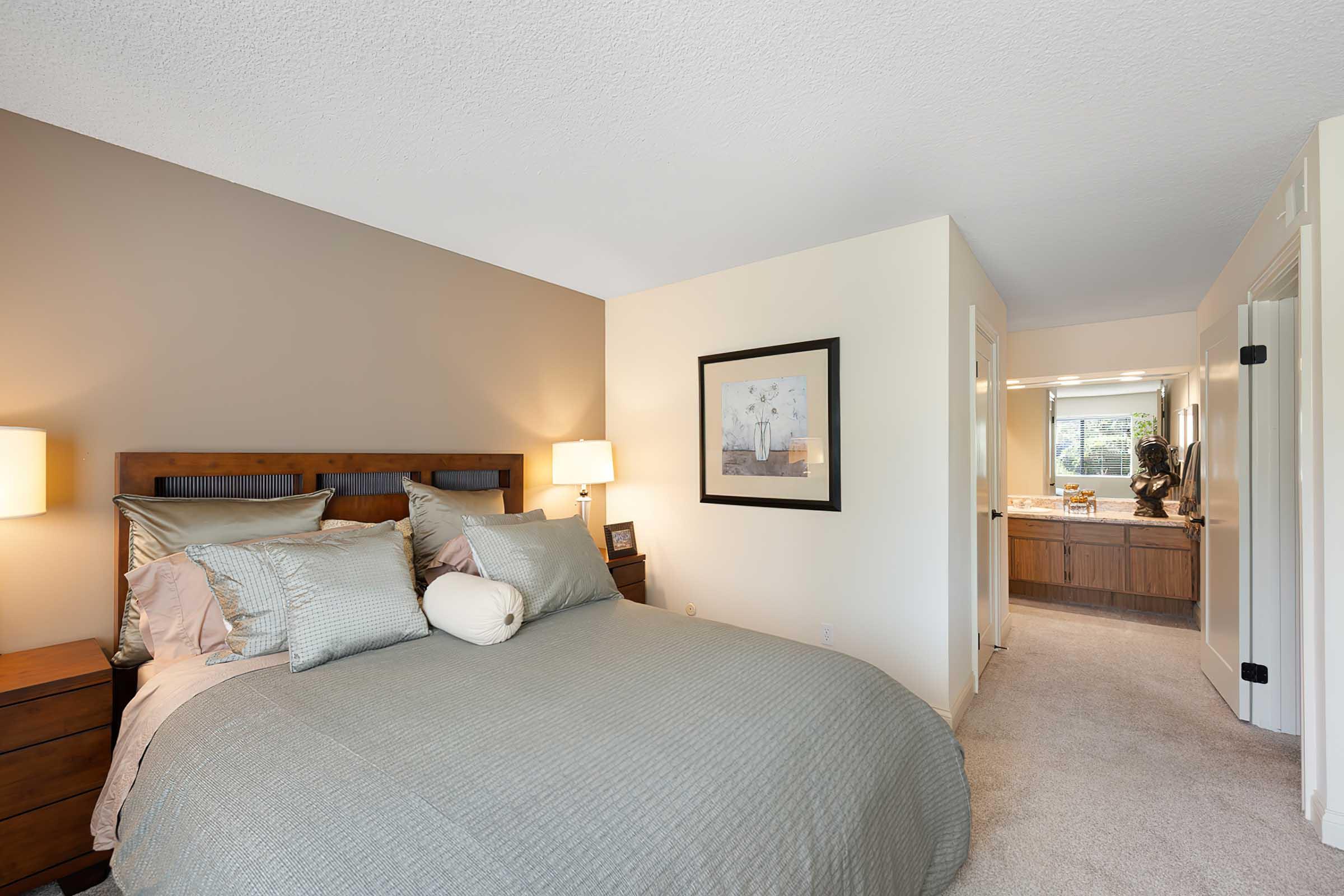 A cozy bedroom featuring a queen-sized bed with light blue bedding, flanked by wooden nightstands with lamps. A piece of artwork hangs on the wall above the bed. The room includes an adjoining bathroom visible through an open door, with modern decor and warm lighting.