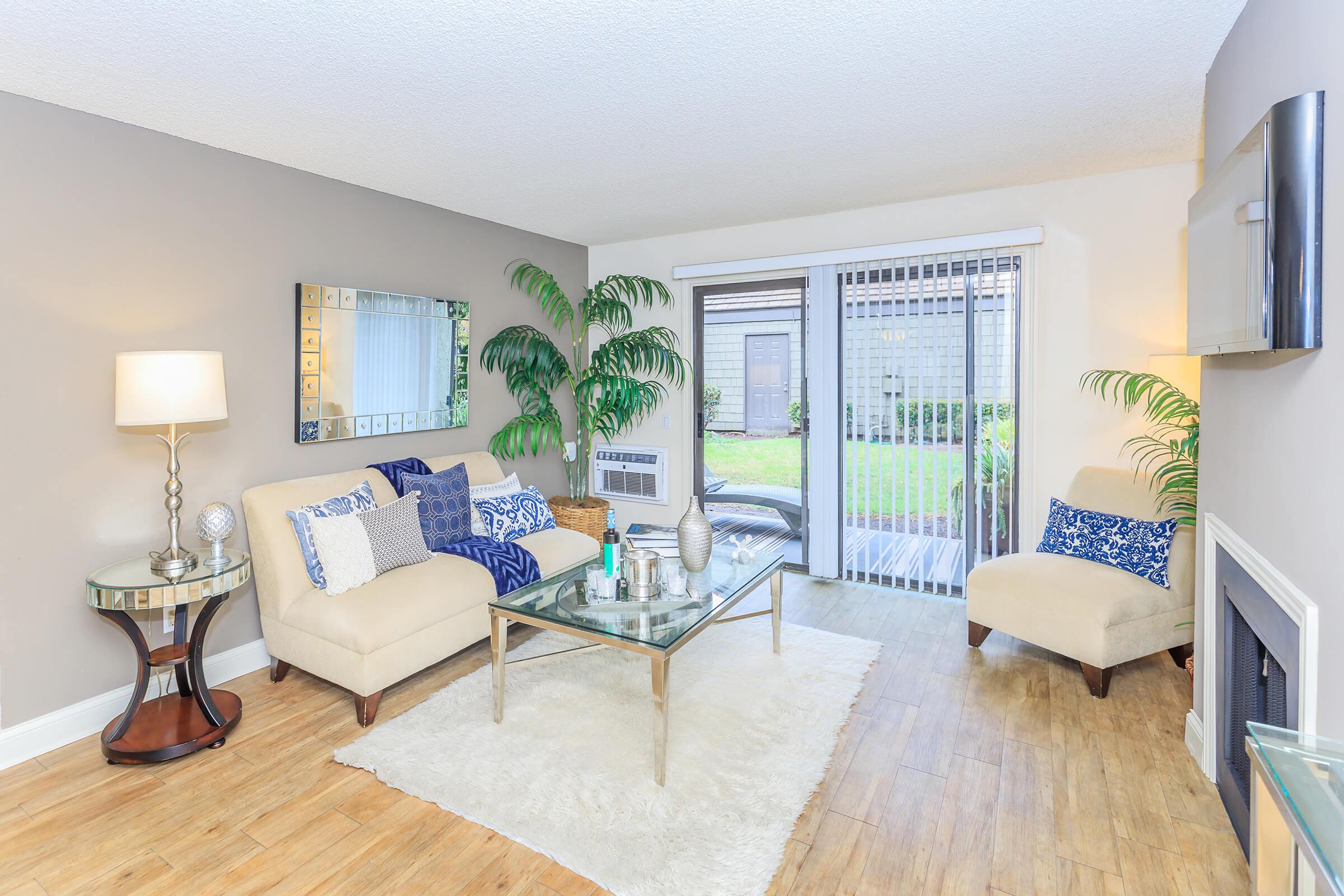 A modern living room featuring a beige couch with blue and white pillows, a glass coffee table on a light rug, a tall lamp, and a large mirror. There's a plant in the corner and a sliding glass door leading to an outdoor space, with light wood flooring throughout the room.