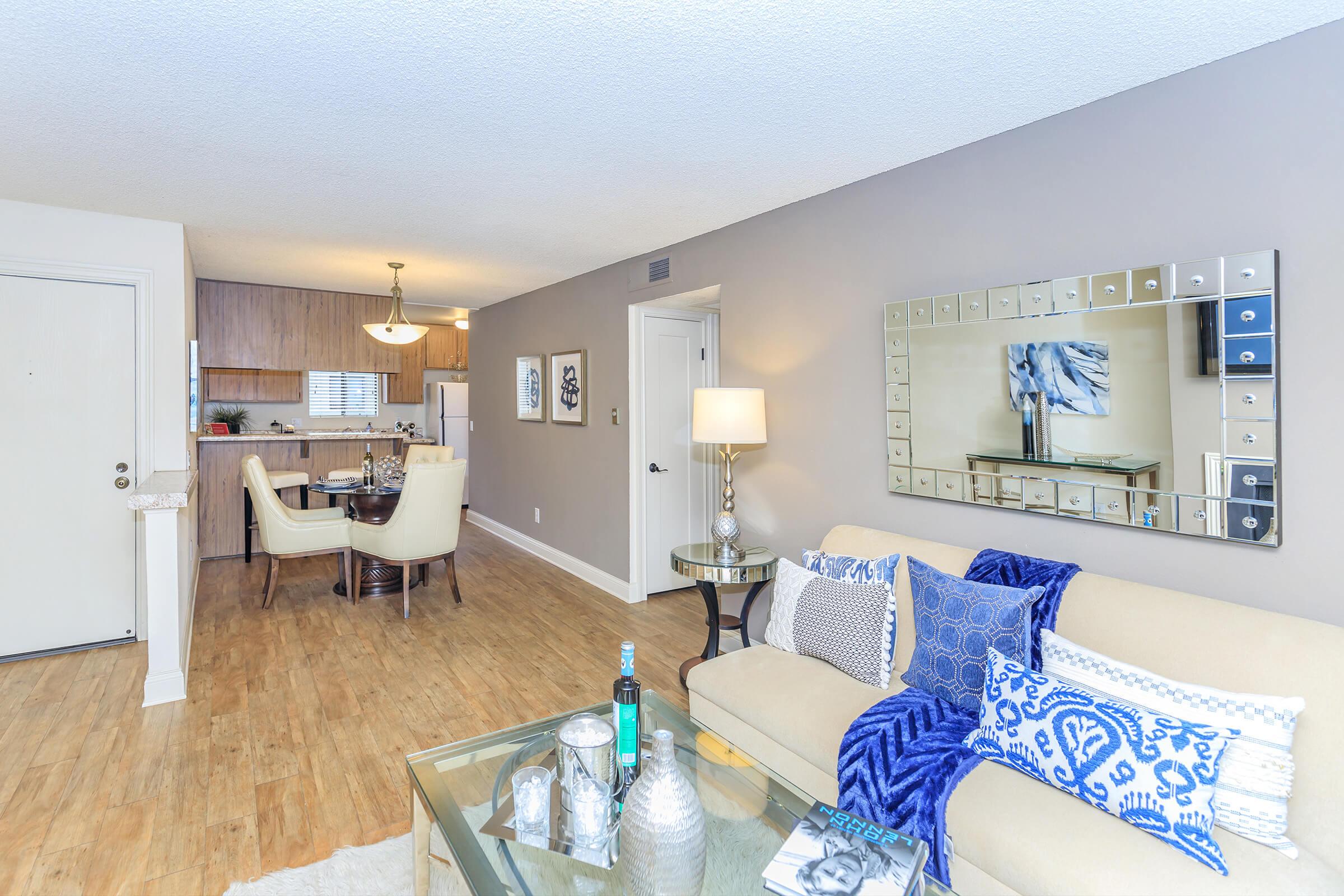 A modern living room featuring a beige sofa with blue and white decorative pillows, a glass coffee table with decorative items, and a large mirror on the wall. There is a dining area in the background with a wooden table and chairs, complemented by warm lighting and tasteful decor.