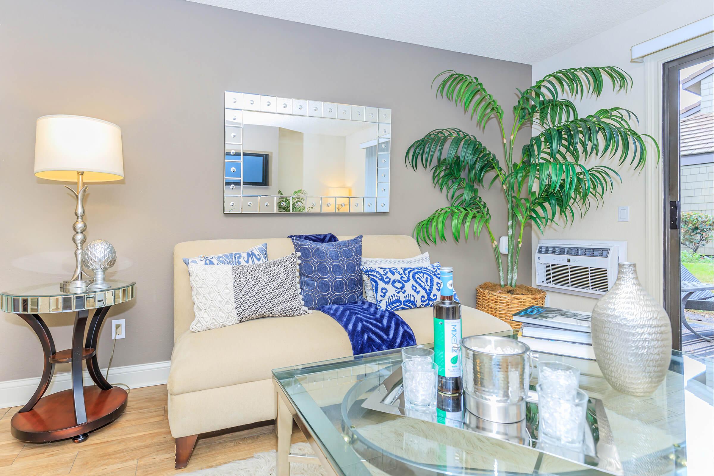 A cozy living room featuring a beige sofa adorned with decorative pillows, a glass coffee table with candles and a decorative bowl, a stylish floor lamp, and a tall potted plant. The space has neutral walls and an air conditioning unit visible in the background, creating a welcoming atmosphere.
