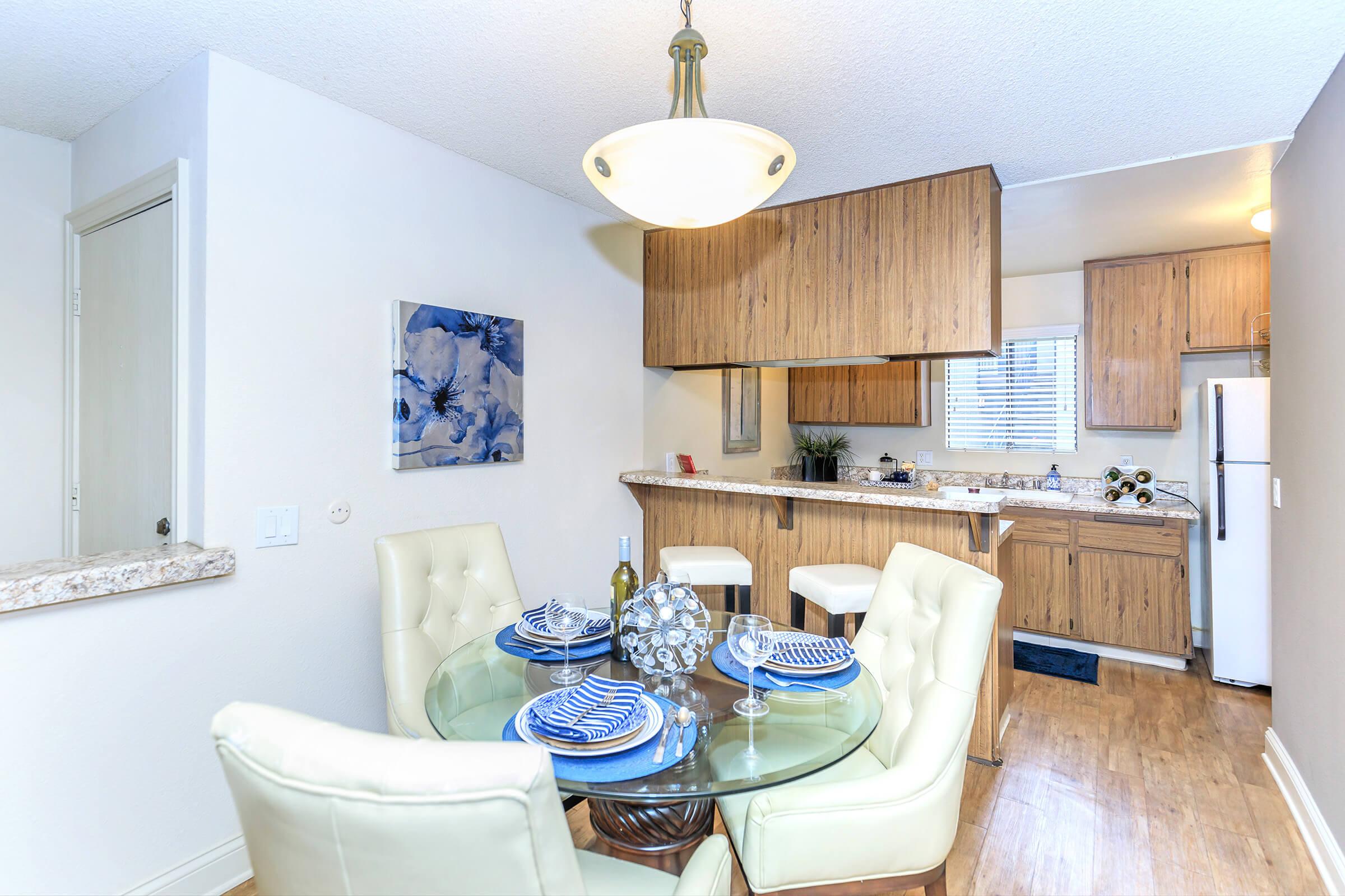 A modern kitchen and dining area featuring a round glass table with white leather chairs, set with blue-patterned dishware. The kitchen has wooden cabinetry and appliances, with a light fixture overhead and an abstract blue artwork on the wall. Natural light enters through a window.