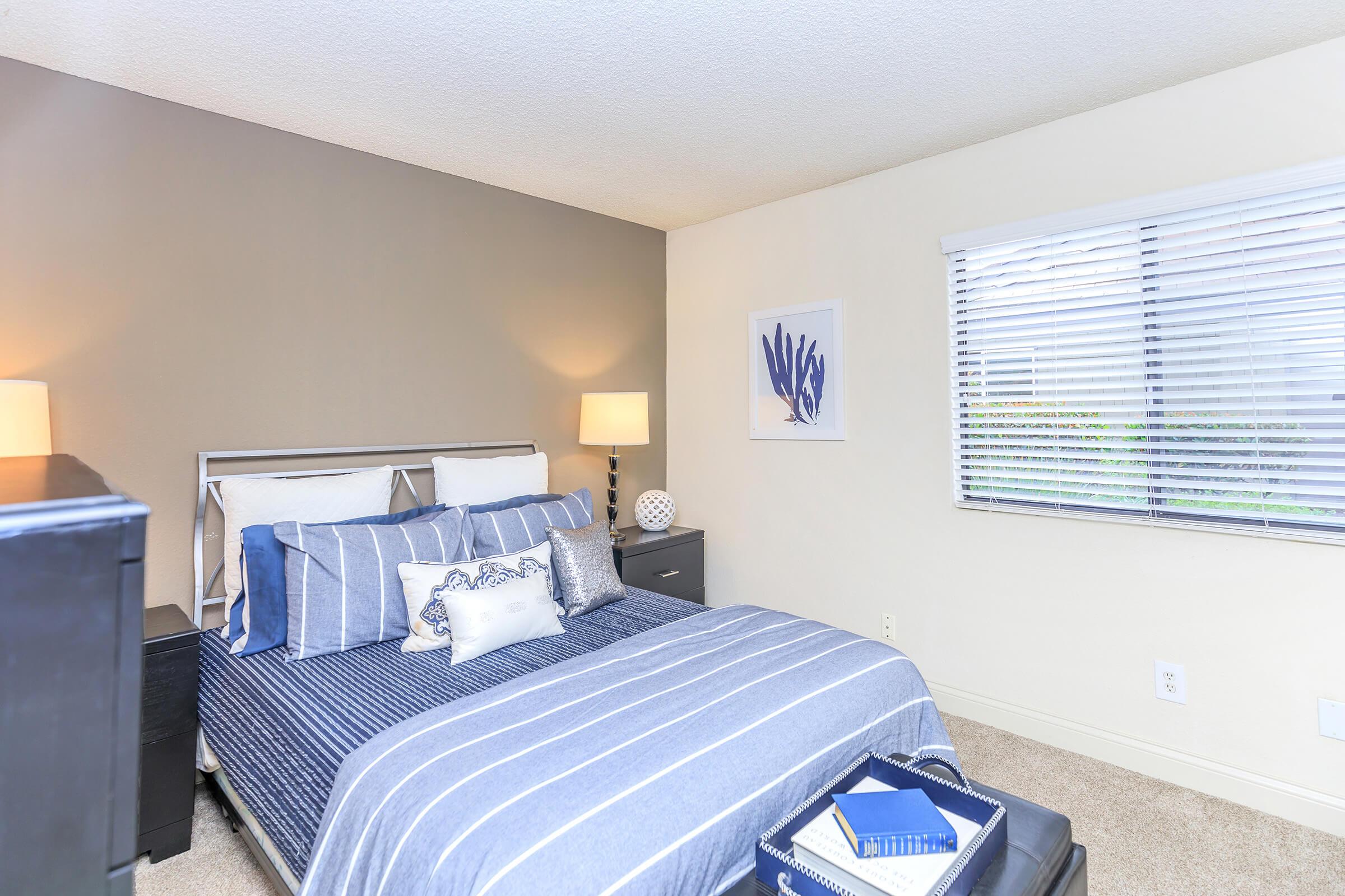 Cozy bedroom featuring a queen-sized bed with blue and white striped bedding, decorative pillows, and matching bedside lamps. A dark wooden dresser is positioned beside the bed. The room has neutral-colored walls and a window with blinds, allowing natural light to enter, complemented by a piece of wall art.