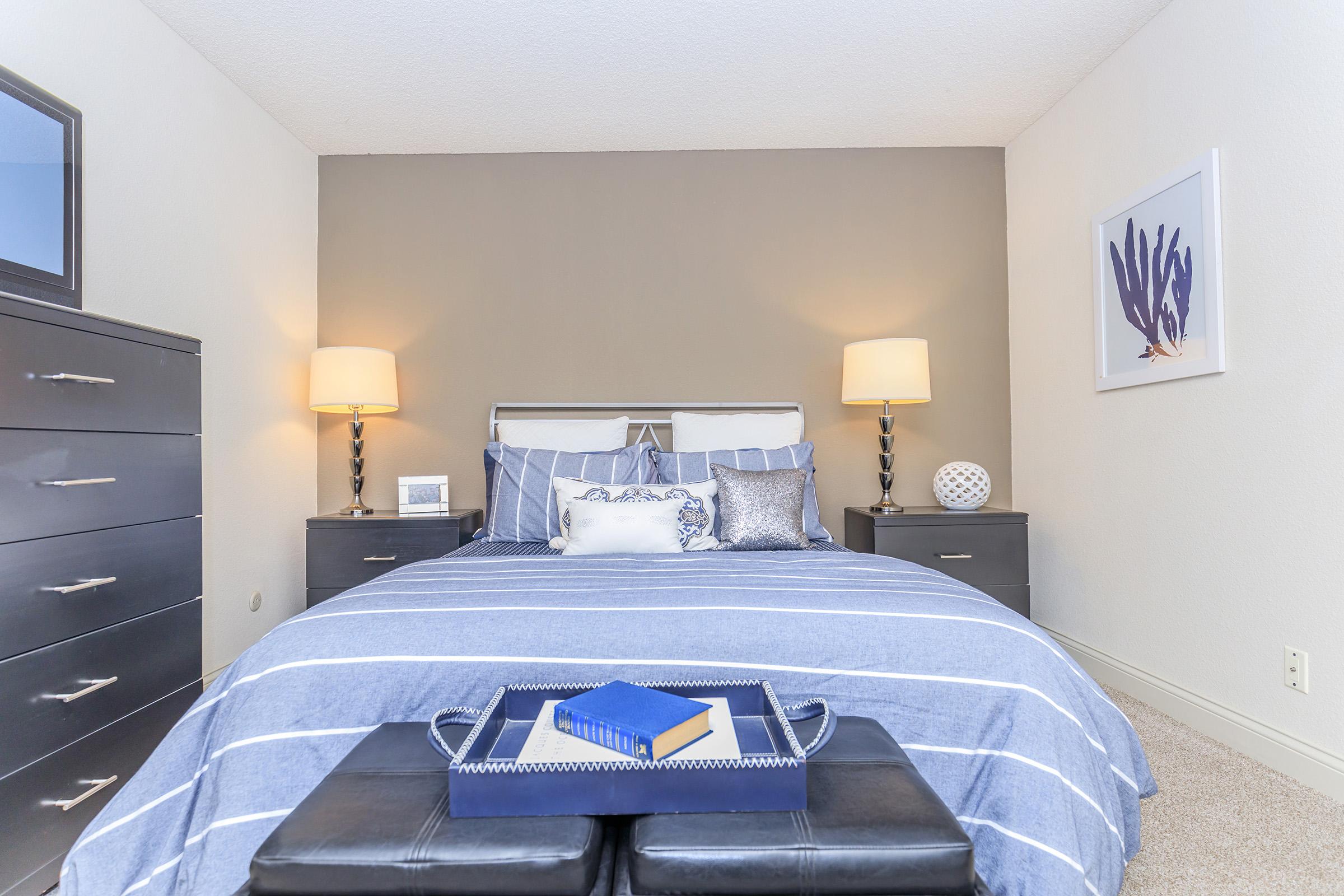 A cozy bedroom featuring a striped blue bedspread, with decorative pillows arranged on the bed. Flanking the bed are two nightstands with lamps. A dark dresser is visible, along with a framed art piece on the wall. A tray with books sits at the foot of the bed, enhancing the inviting atmosphere.