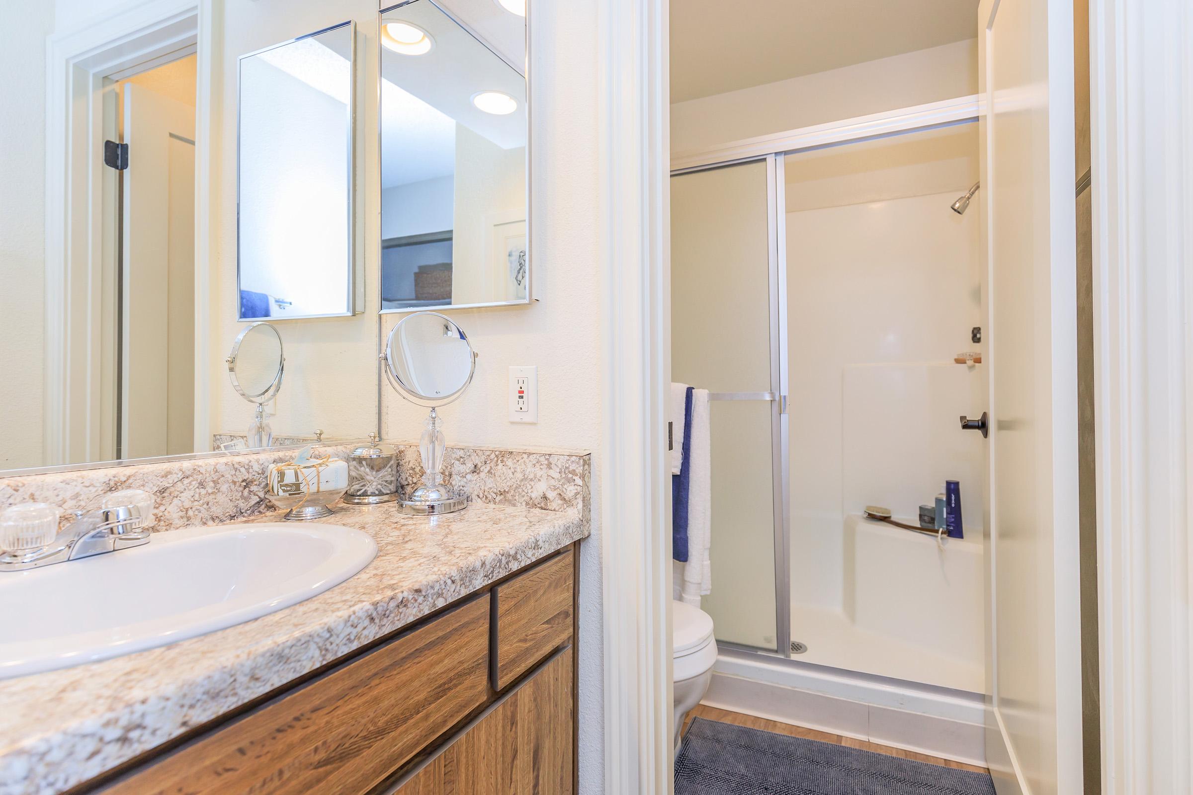 A clean bathroom featuring a marble countertop with a sink, two round mirrors, and wooden cabinetry. To the right, there's a shower with a clear glass door and a tiled floor. There are toiletries visible on the shower ledge and a towel hanging nearby. Overall, the space is well-lit and tidy.