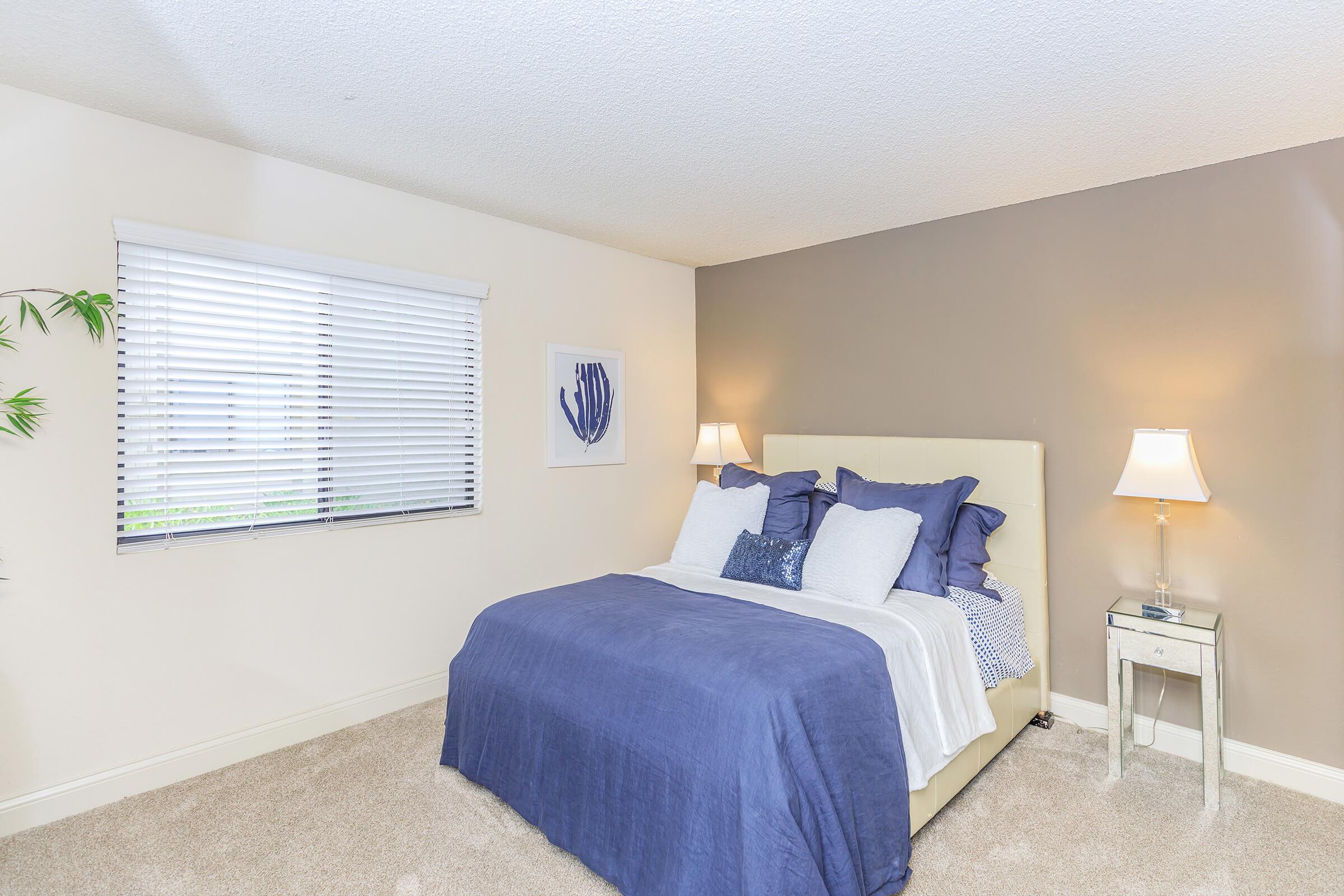 A cozy bedroom featuring a light-colored bed with blue and white bedding, flanked by two bedside lamps. The walls are a soft brown and beige, with a window adorned with blinds. A framed artwork of a coral print is mounted next to the window, adding a touch of decor. The floor is carpeted in a neutral tone.