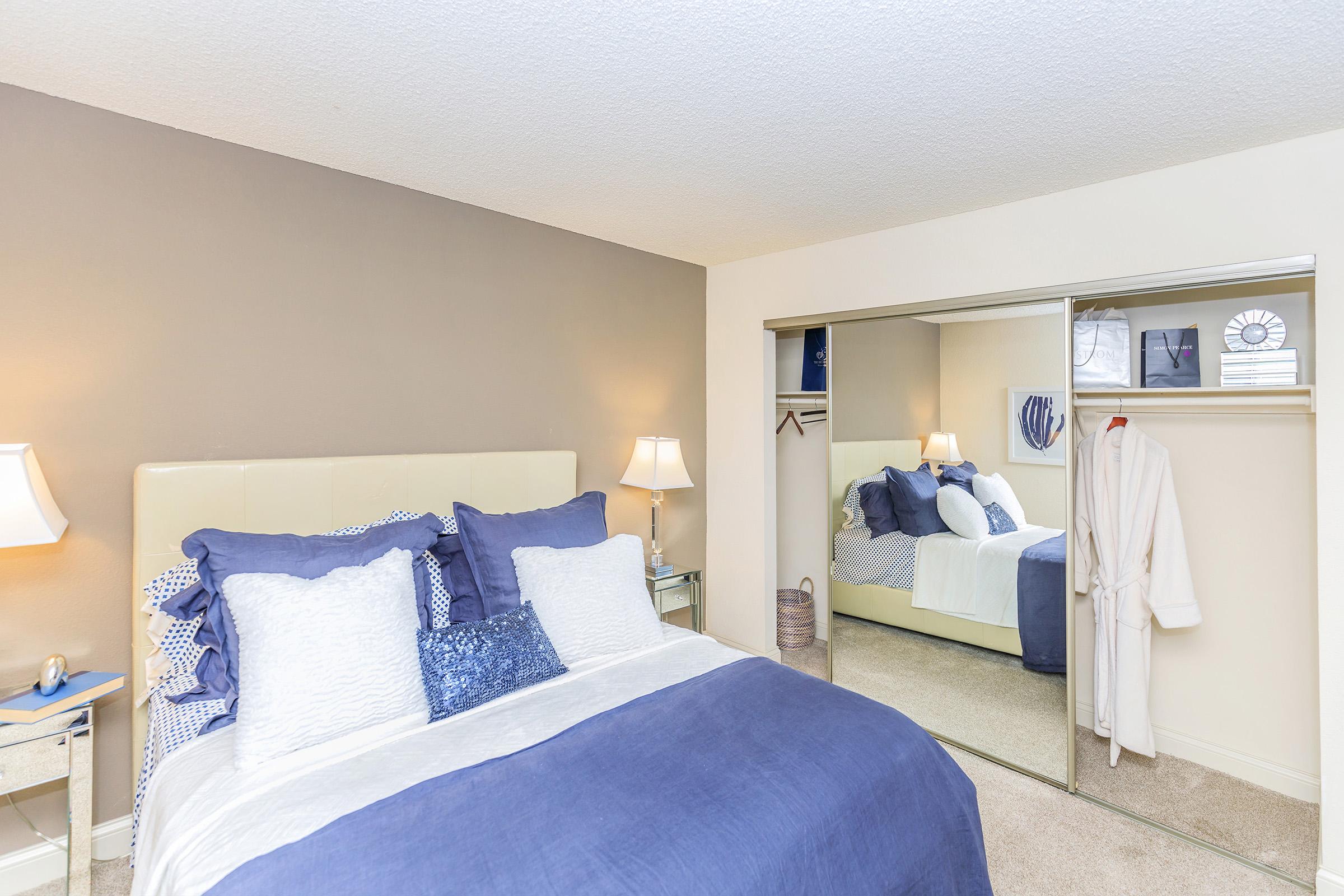 A cozy bedroom featuring a king-sized bed with blue and white bedding, two bedside lamps, and a mirrored closet. A plush white robe hangs in the closet, and a light-colored carpet complements the decor. The walls are painted in neutral tones, creating a relaxing atmosphere.
