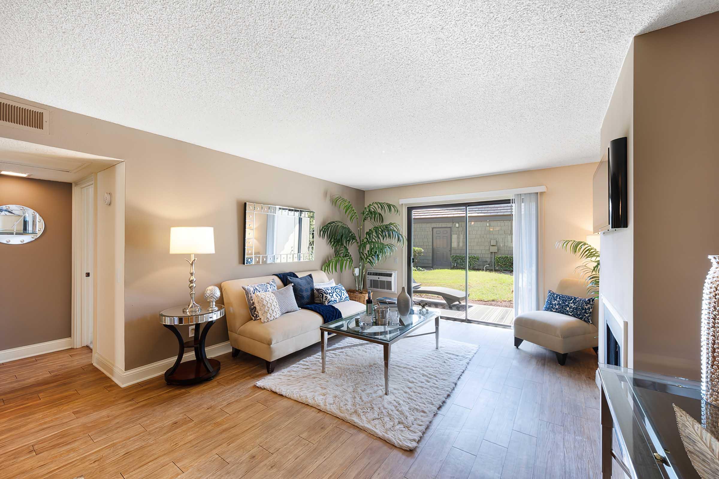 A modern living room featuring a beige sofa with decorative pillows, a glass coffee table on a soft rug, a stylish side table with a lamp, and large windows letting in natural light. Green plants add a touch of nature, and there’s a view of a patio outside. The walls are painted in a warm, neutral tone.