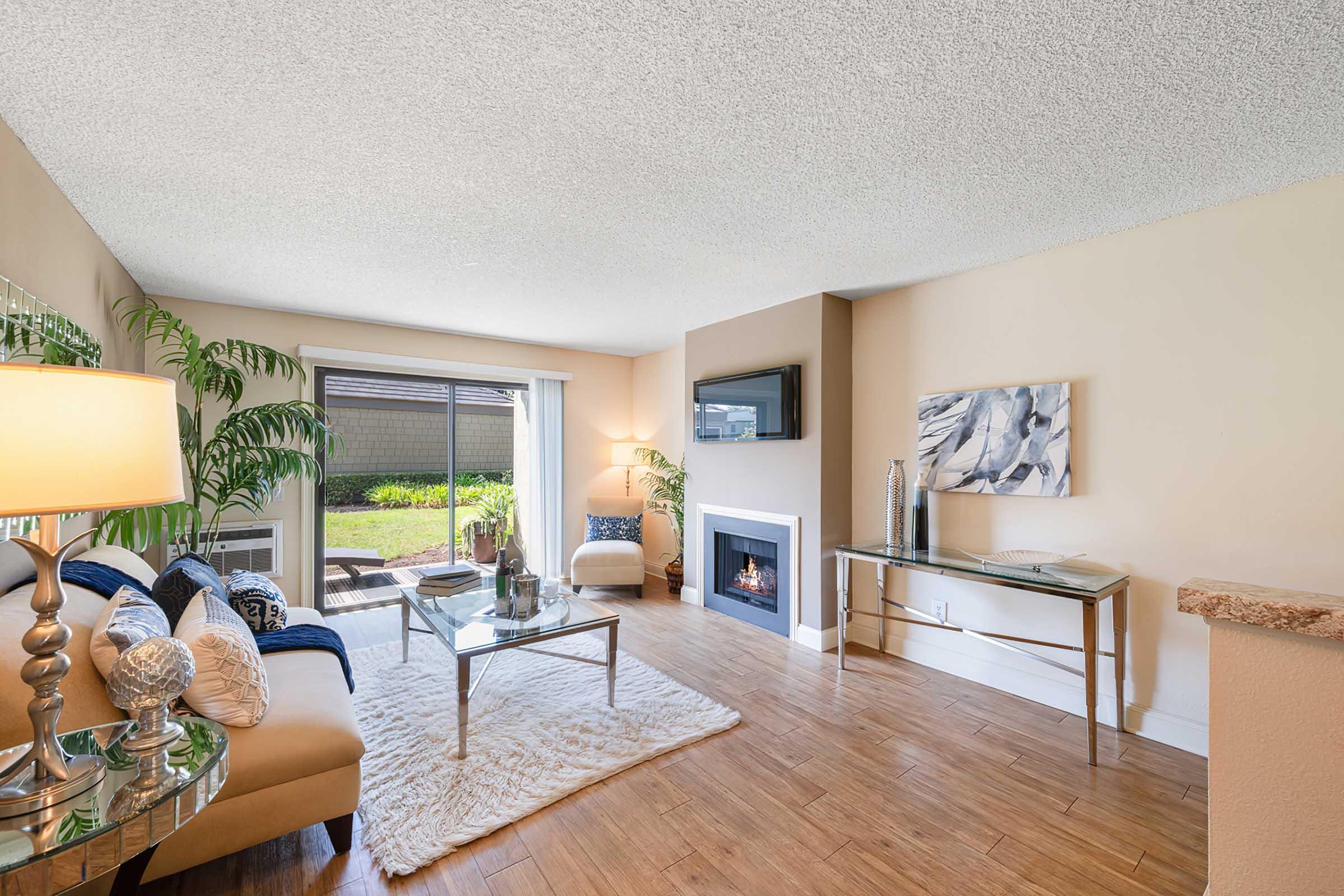 Bright and inviting living room featuring a beige couch with blue accent pillows, a glass coffee table, and a cozy fireplace. Large windows allow natural light to fill the space, complemented by indoor plants and modern decor. A plush rug adds warmth to the wooden flooring.