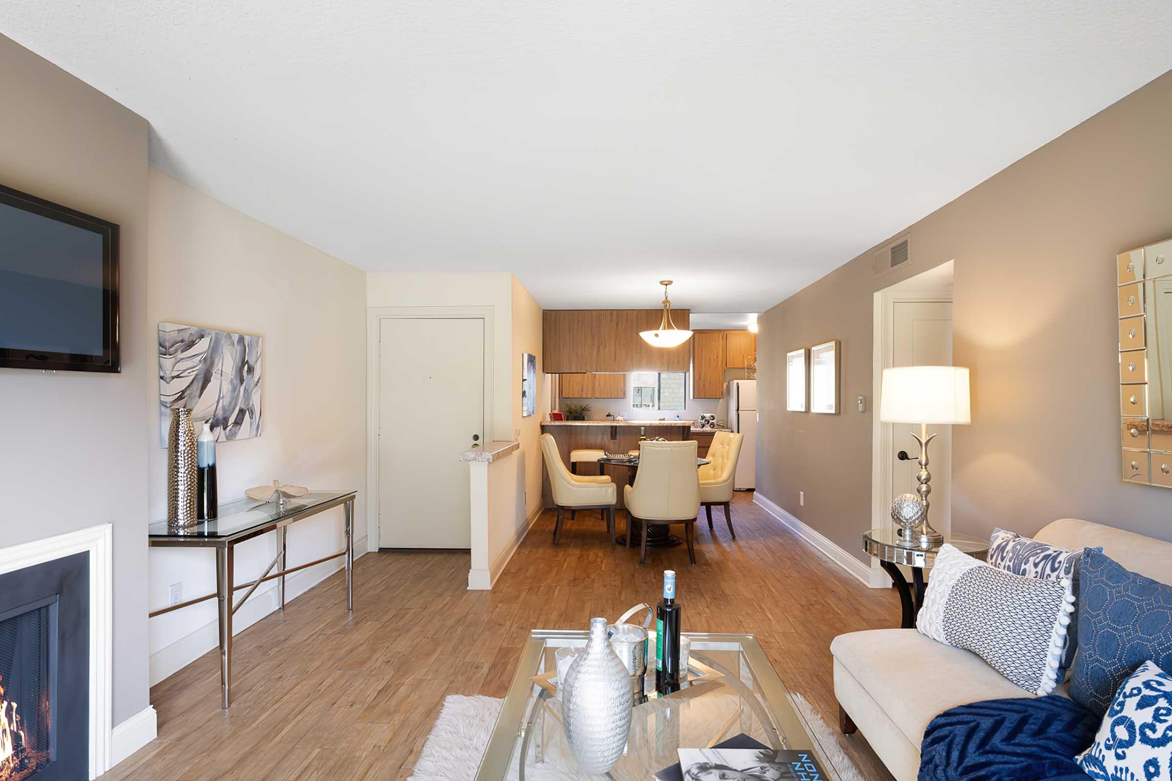 A modern living room featuring a beige couch with decorative pillows, a glass coffee table with bottles and a candle. A flat-screen TV is mounted on the wall. The room leads into an open kitchen and dining area with light wooden cabinetry and a round dining table with upholstered chairs. Natural light fills the space.