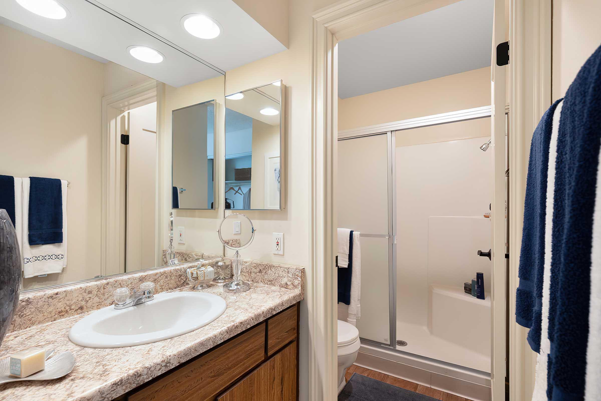 A well-lit bathroom featuring a marble countertop with a sink, a large mirror, and a vanity area. On the right, there is a shower stall with a glass door, and towels hanging on a rack. Neutral-colored walls and wooden flooring complete the tidy and modern aesthetic.