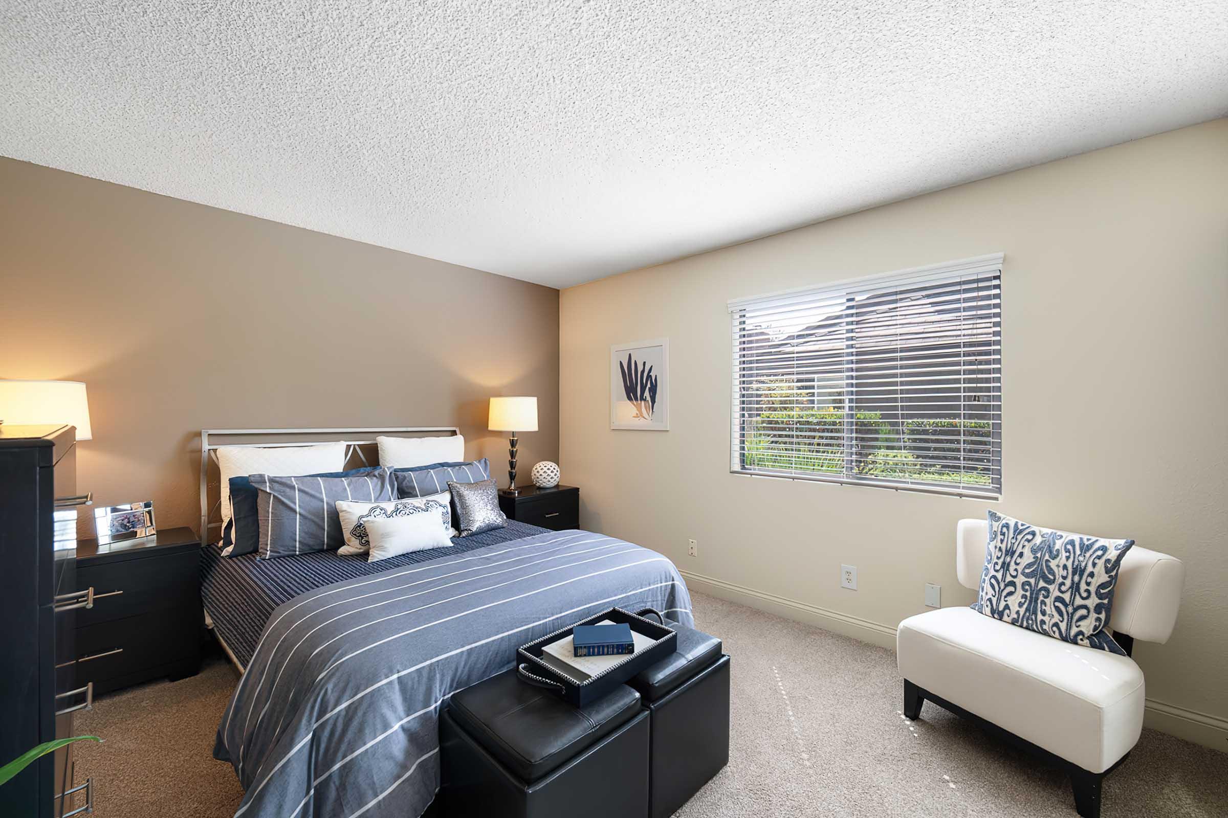 A cozy bedroom featuring a queen-sized bed with blue striped bedding, two bedside lamps, and a black storage ottoman. A white armchair is positioned next to the bed. The room has neutral-colored walls and a window with blinds, allowing natural light to fill the space, with a view of greenery outside.